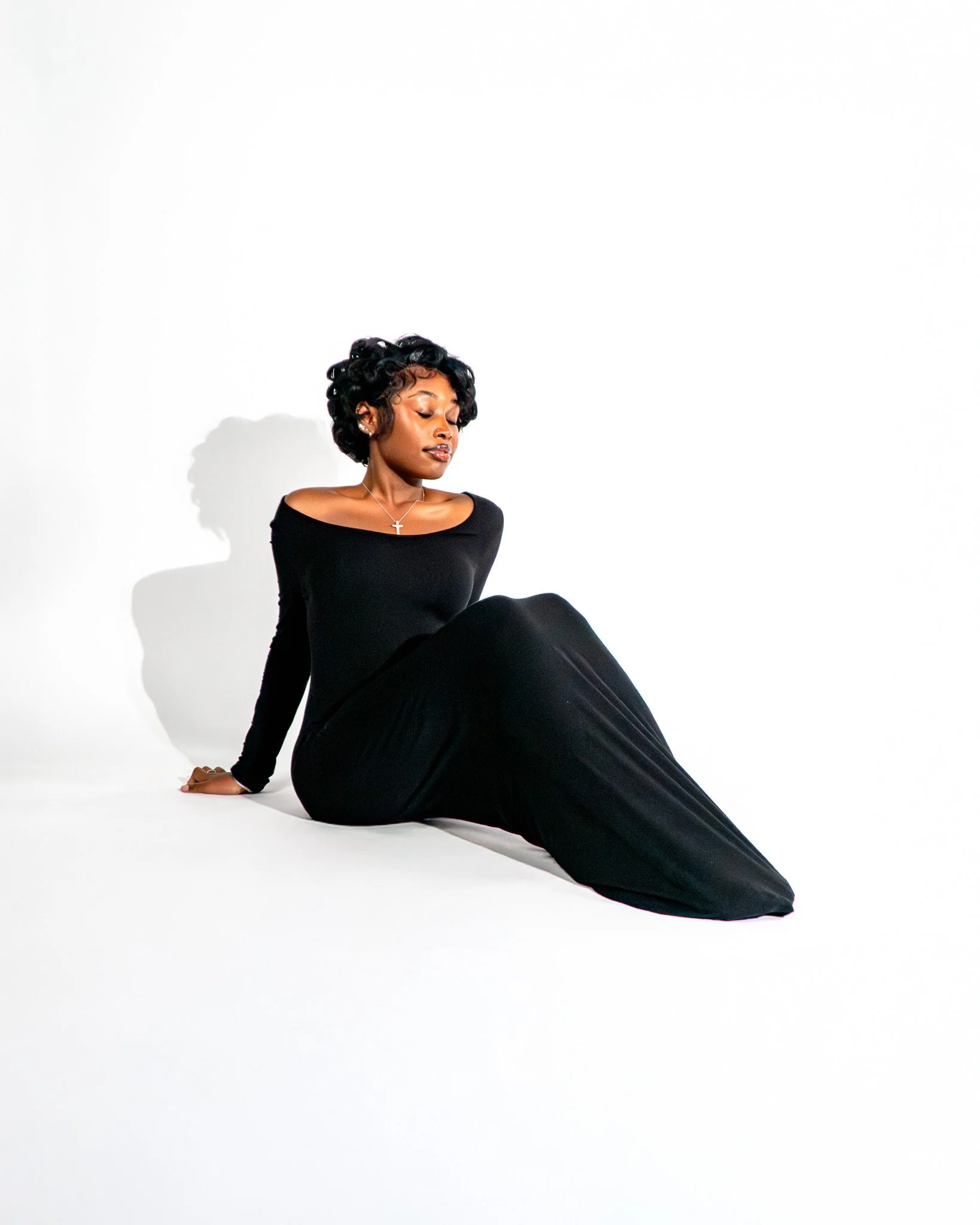 A woman with short, curly black hair, wearing a black off-shoulder dress, is sitting on the floor against a white background. She has her eyes closed, head tilted slightly downward, and a calm expression. She is accessorized with a silver cross necklace and small earrings, with a shadow cast on the white wall behind her.