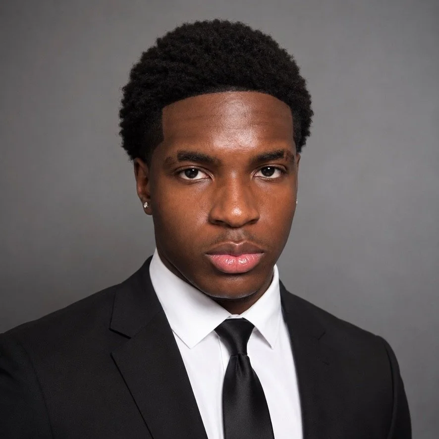 Portrait of a young Black man in a black suit, white shirt, and black tie against a gray background.