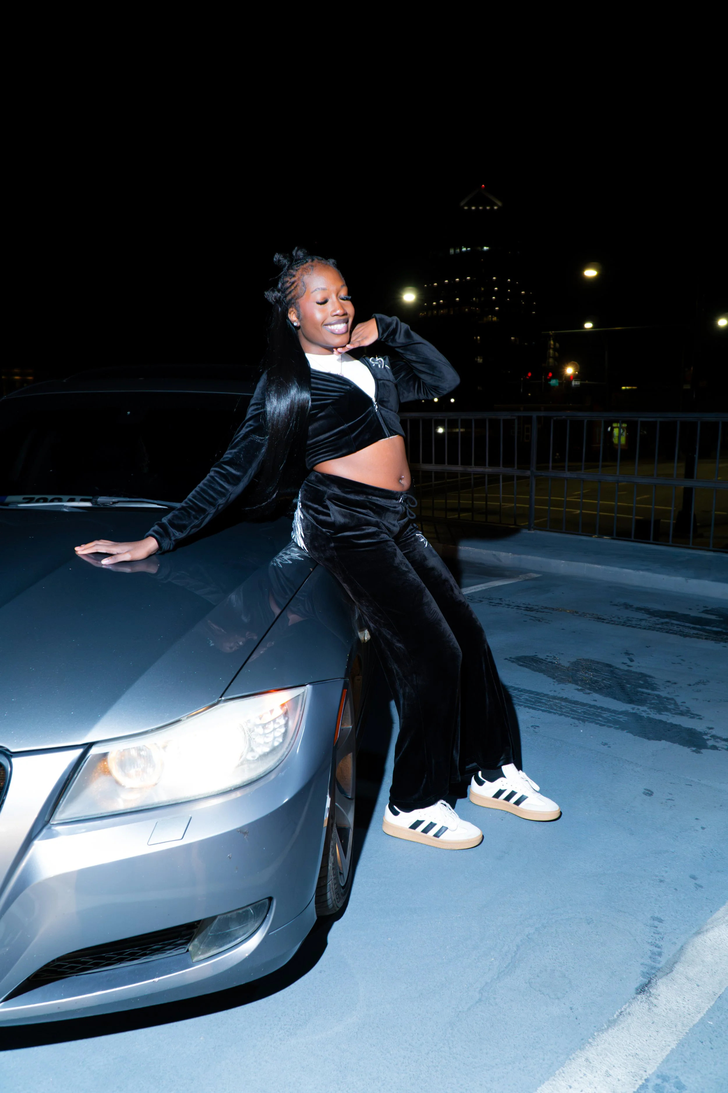 A young woman with dark skin, long black hair, and a belly piercing, standing on a car hood at night in a parking lot. She is smiling, wearing a black velour tracksuit, white shirt, and Adidas sneakers, with a city skyline and streetlights in the background.