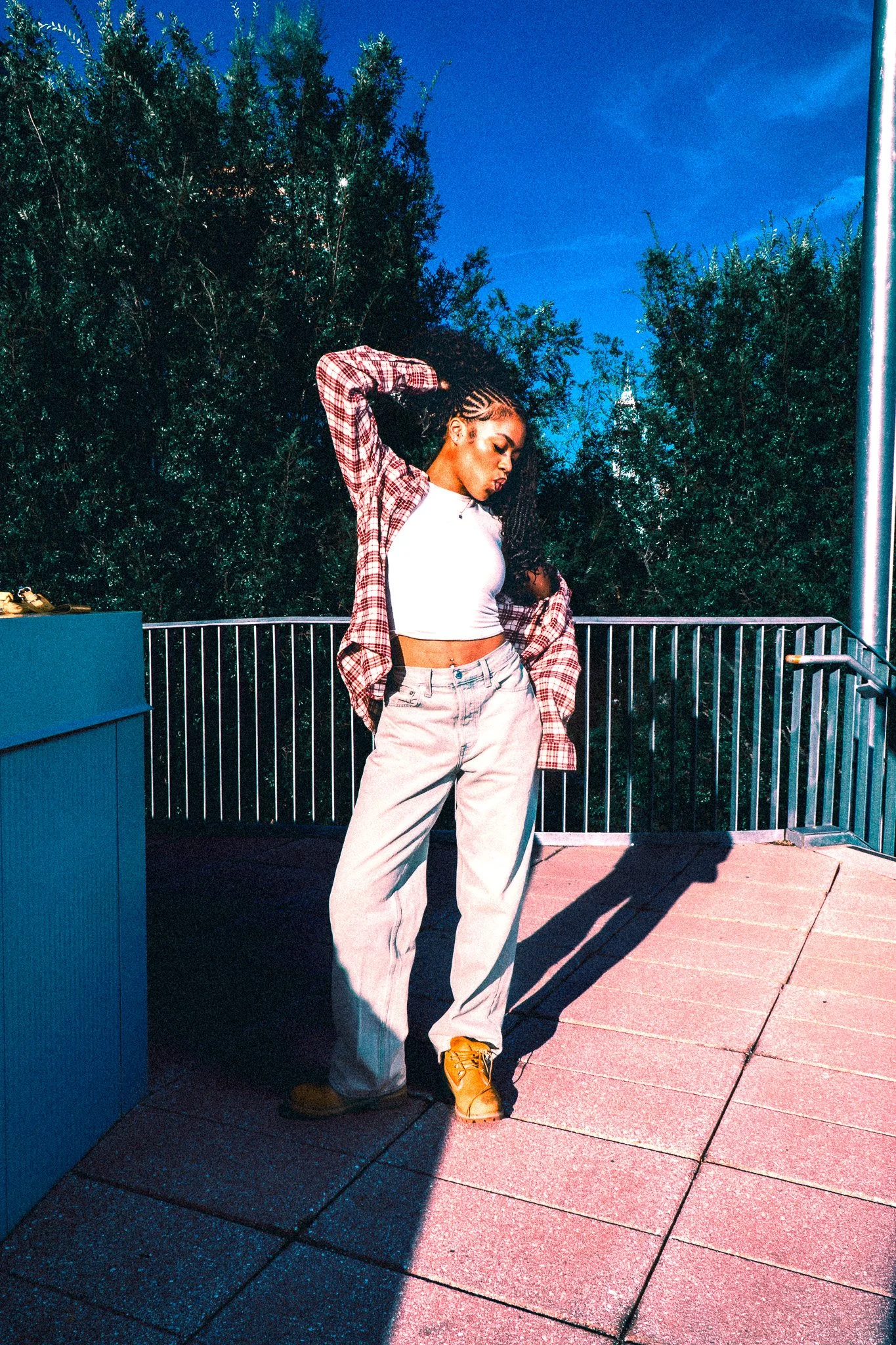A young woman poses on a balcony during early evening, wearing a white crop top, baggy beige pants, a red plaid shirt, and tan boots, with braided hair, standing with one hand behind her head against a backdrop of green trees and a bright blue sky.