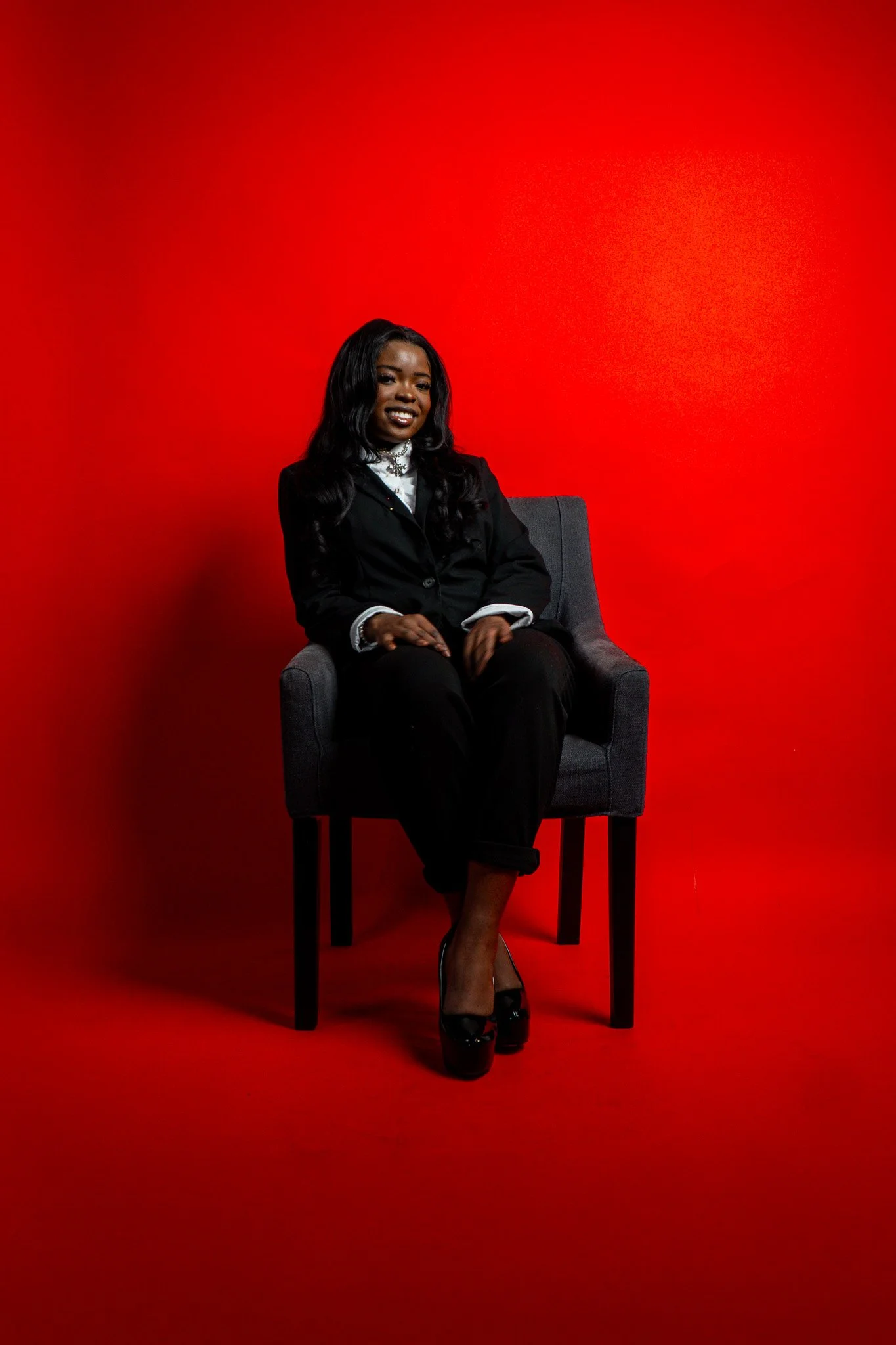 A woman with long black hair, dressed in a black outfit with a white shirt, sitting on a gray chair against a red background.