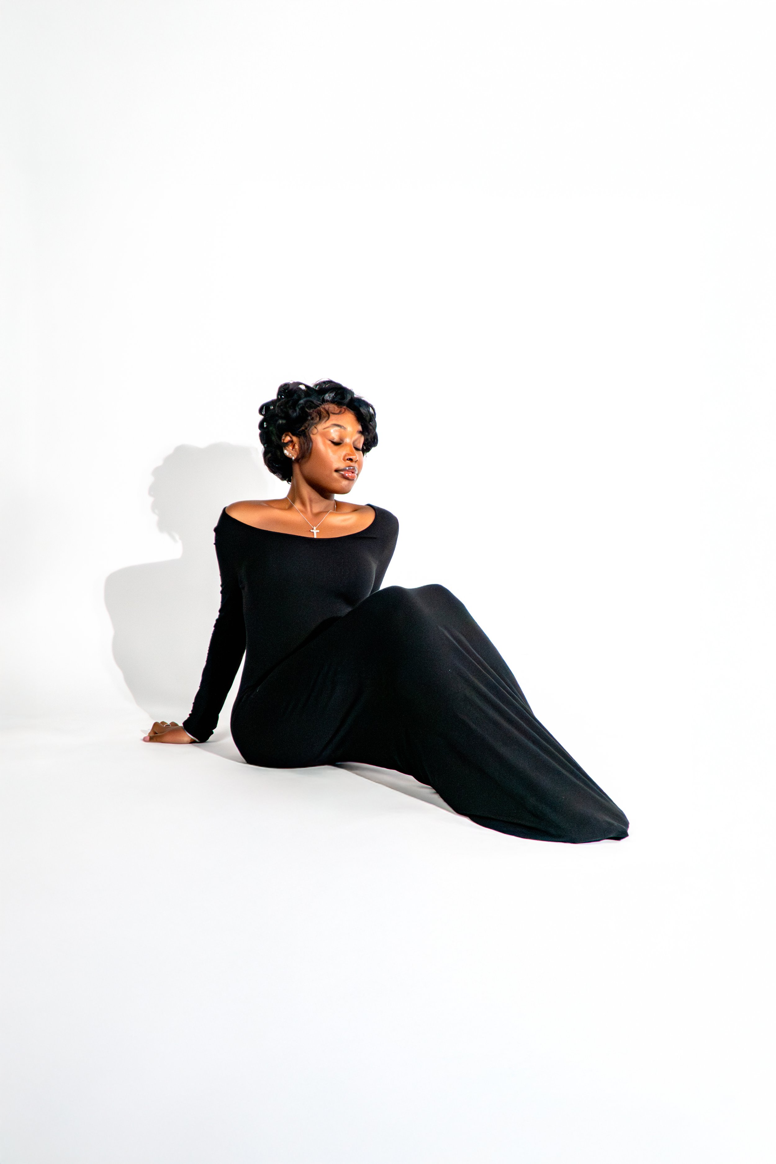 A woman sitting on the floor against a plain white wall, wearing a black long-sleeve dress and showing a calm, contemplative expression.