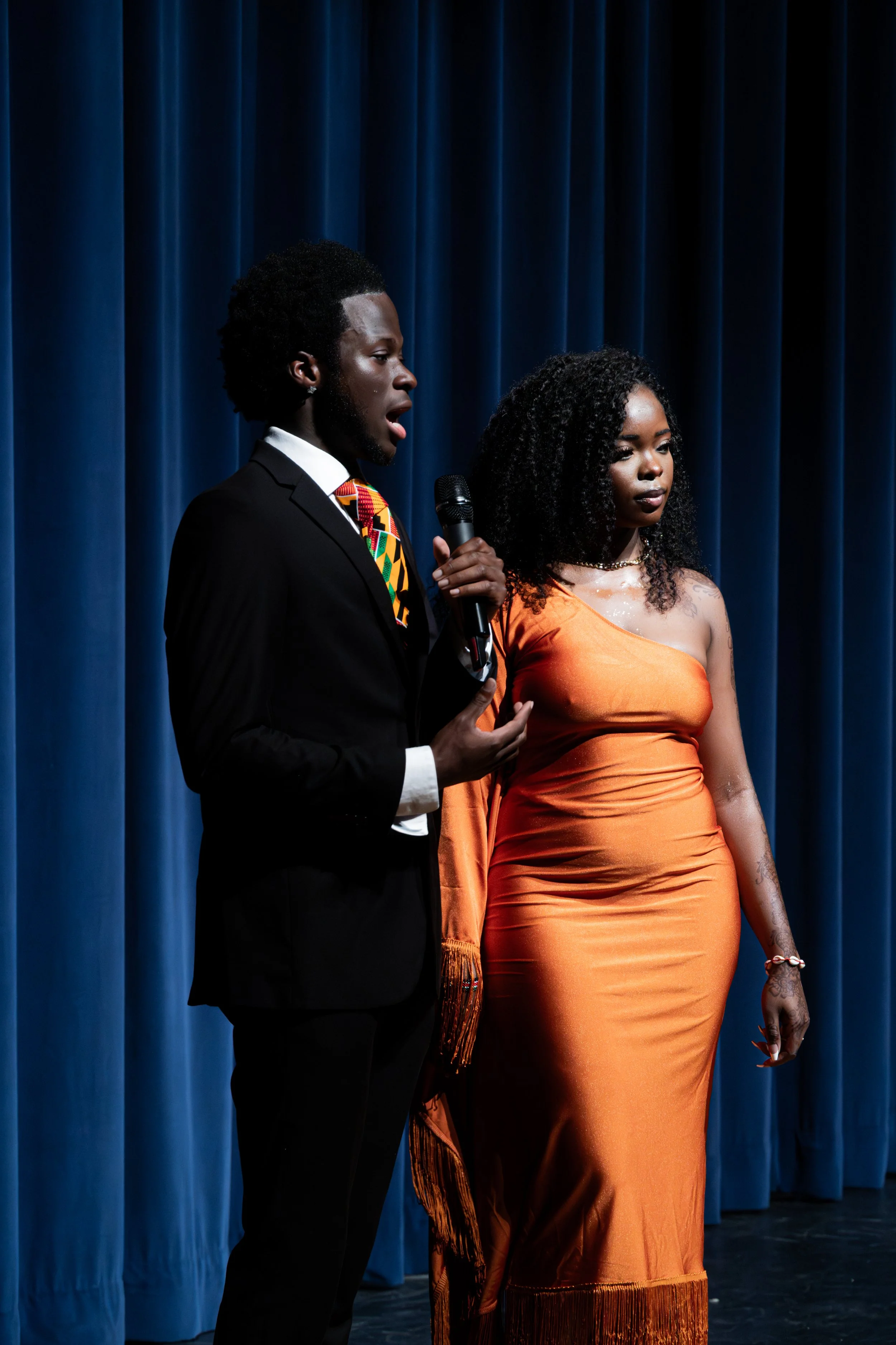 A man in a black suit with a colorful patterned tie and a woman in an orange dress on stage in front of blue curtains, with the man holding a microphone and speaking, while the woman listens.
