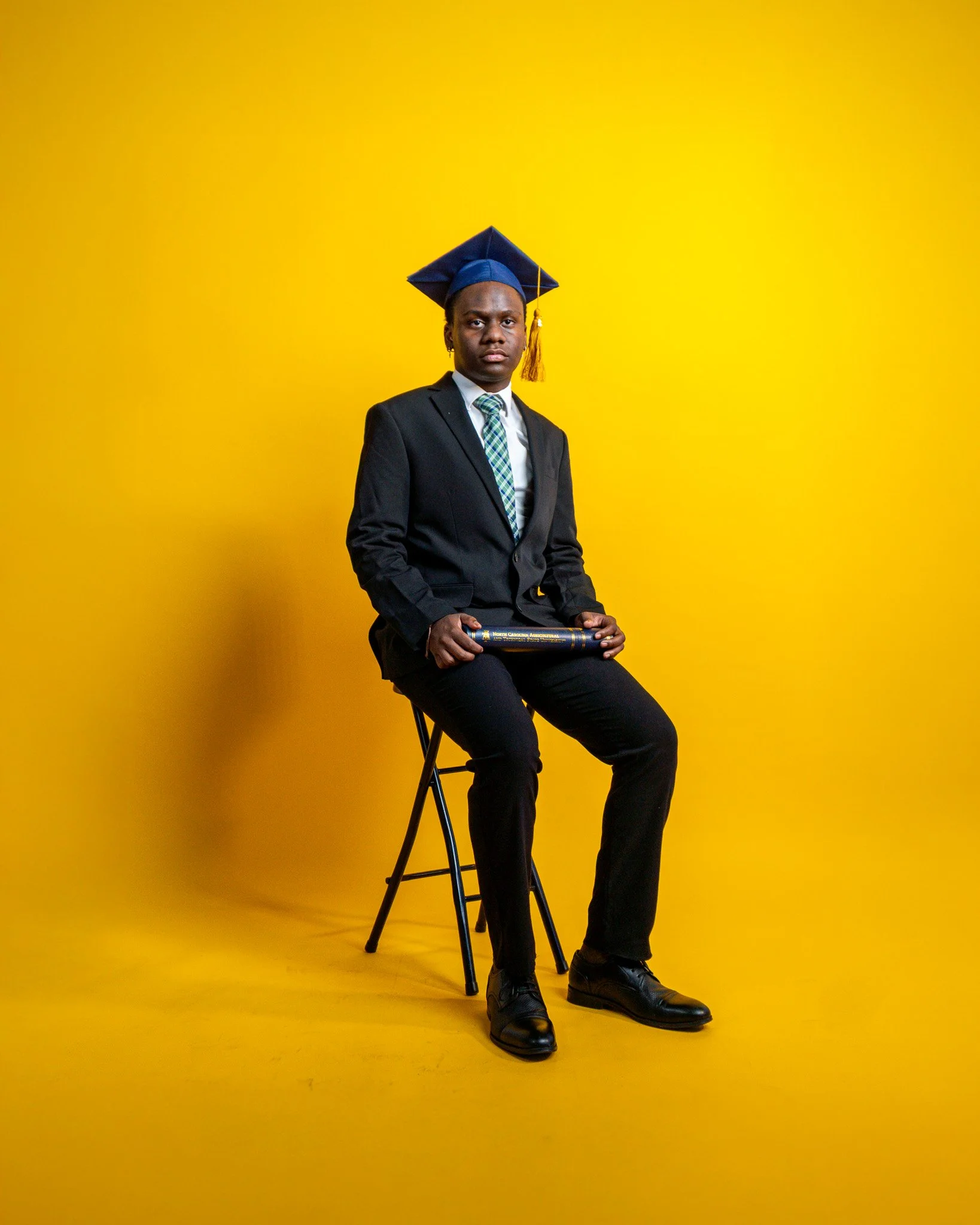 A young man in a black suit, white shirt, and patterned tie is sitting on a black chair against a bright yellow background. He is holding a diploma or certificate in his lap and wearing a blue graduation cap.