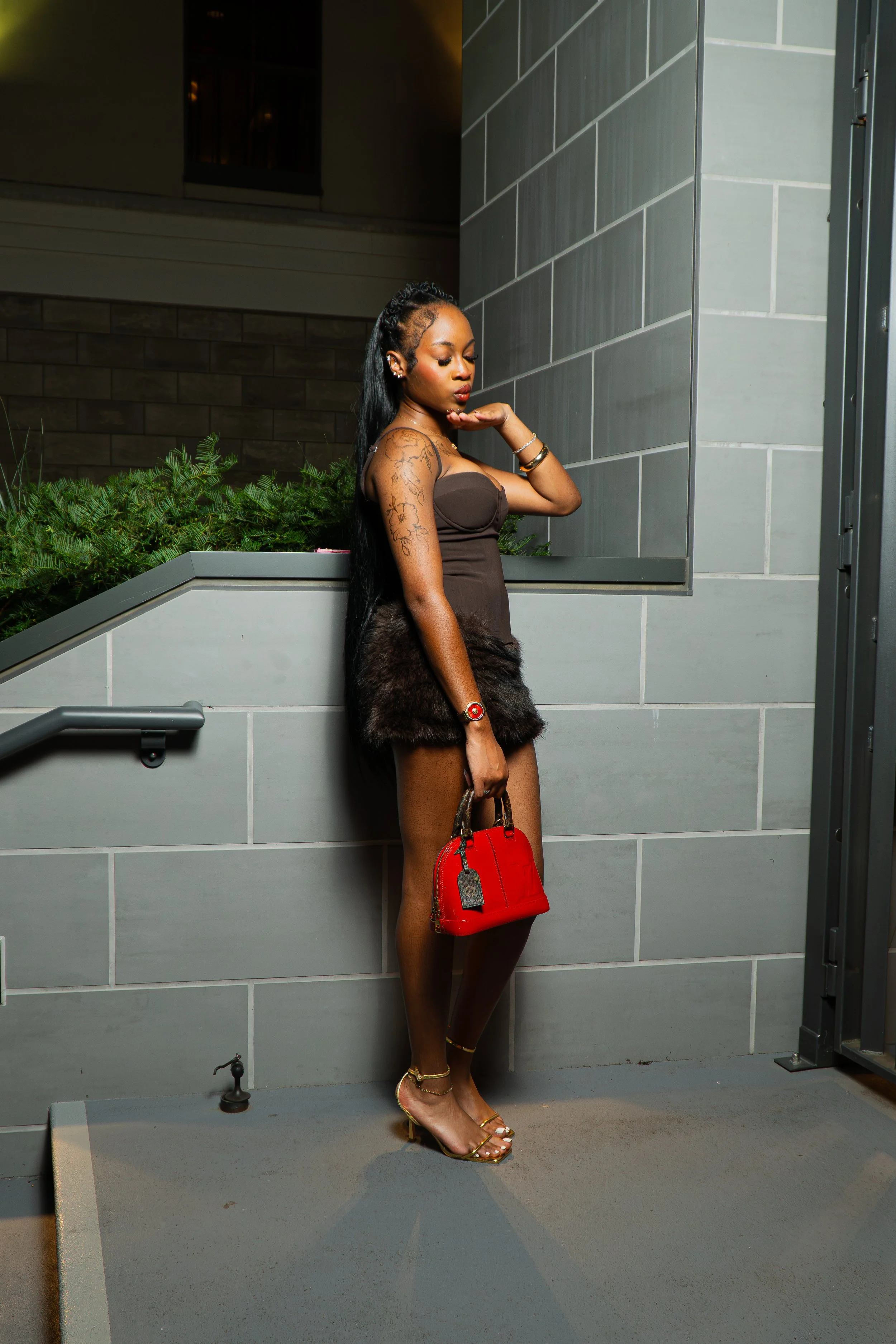 A woman with long black hair, wearing a strapless brown dress with fur at the bottom, gold high heels, and gold jewelry, stands against a gray tiled outdoor wall at night, holding a red purse and striking a pose with her eyes closed and one hand near her face.