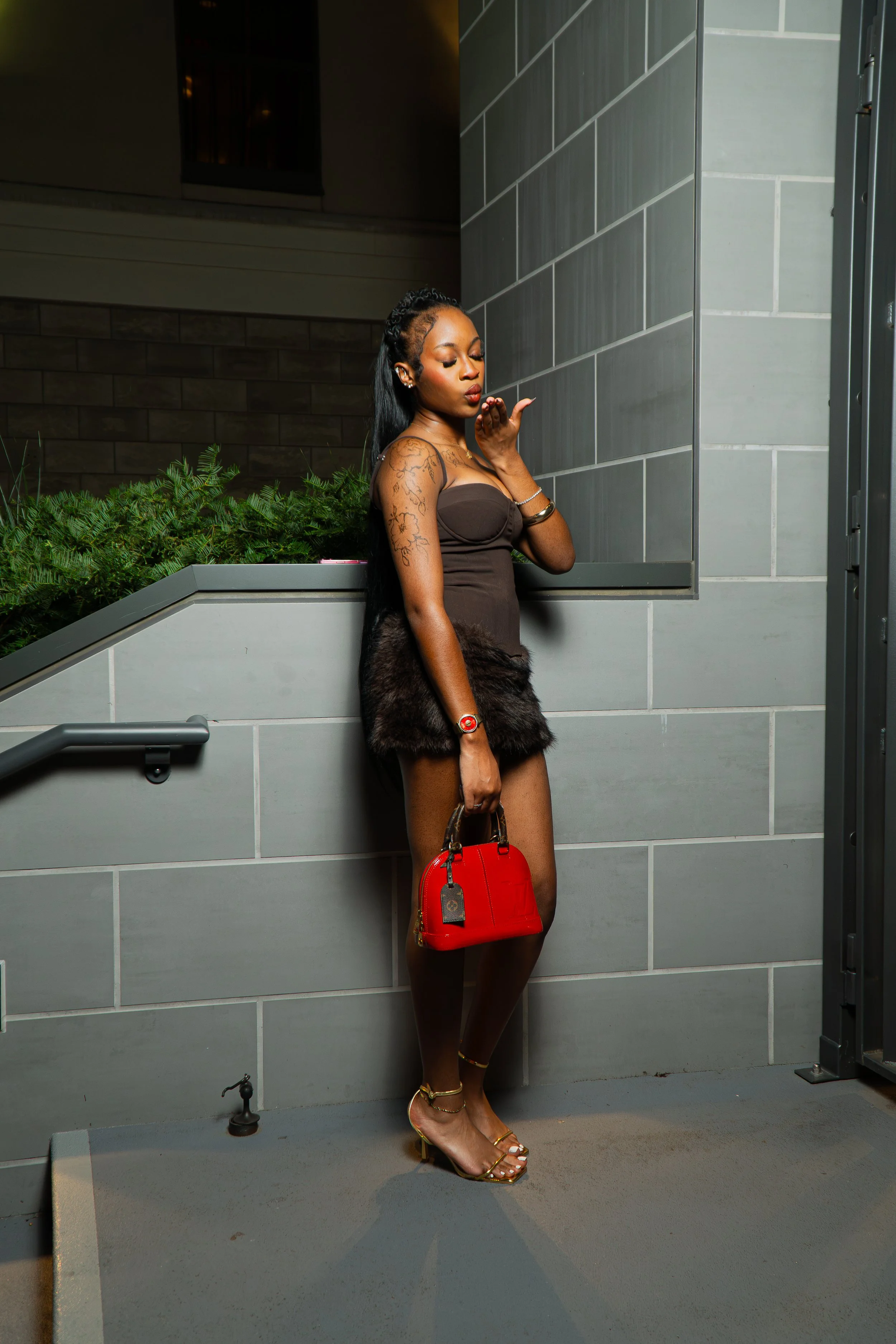 A woman with long black hair, wearing golden high heels, a black strapless dress with fur trim, and gold accessories, looks down and blows a kiss, holding a red handbag, against a background of gray tiled walls and greenery.