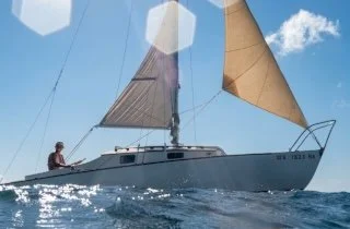 A sailboat with two beige sails on the ocean under a blue sky with white clouds, with a person sitting on the deck.