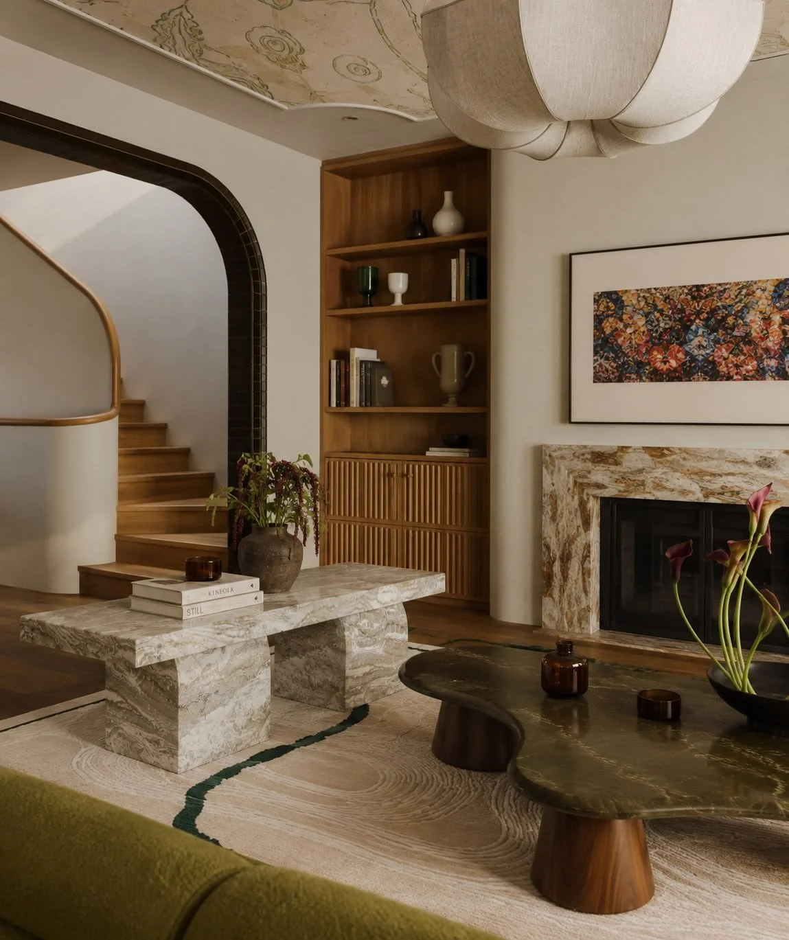 Living room with a marble coffee table, a green sofa, a fireplace with a marble surround, a wooden bookshelf, a staircase, and a large fabric-covered pendant light.
