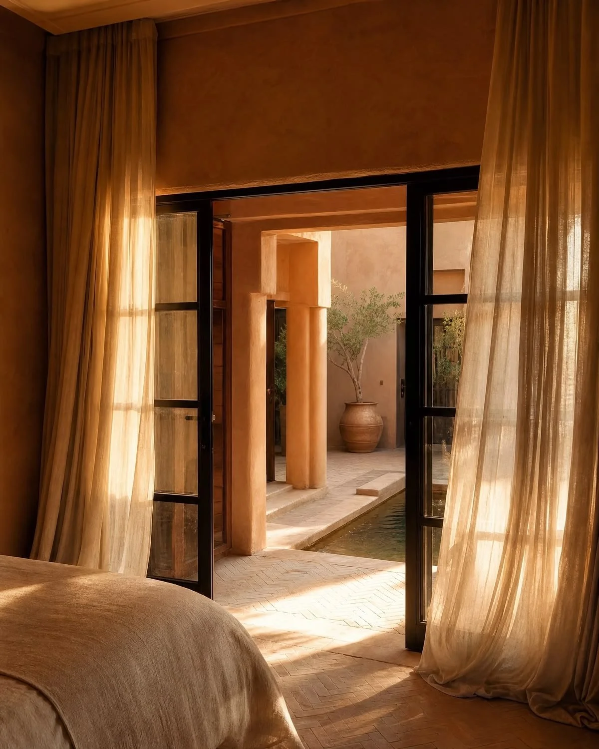 View from inside a room looking through open black-framed glass doors to an outdoor courtyard with beige walls, columns, a potted plant, and a small water feature, with sheer beige curtains framing the scene.