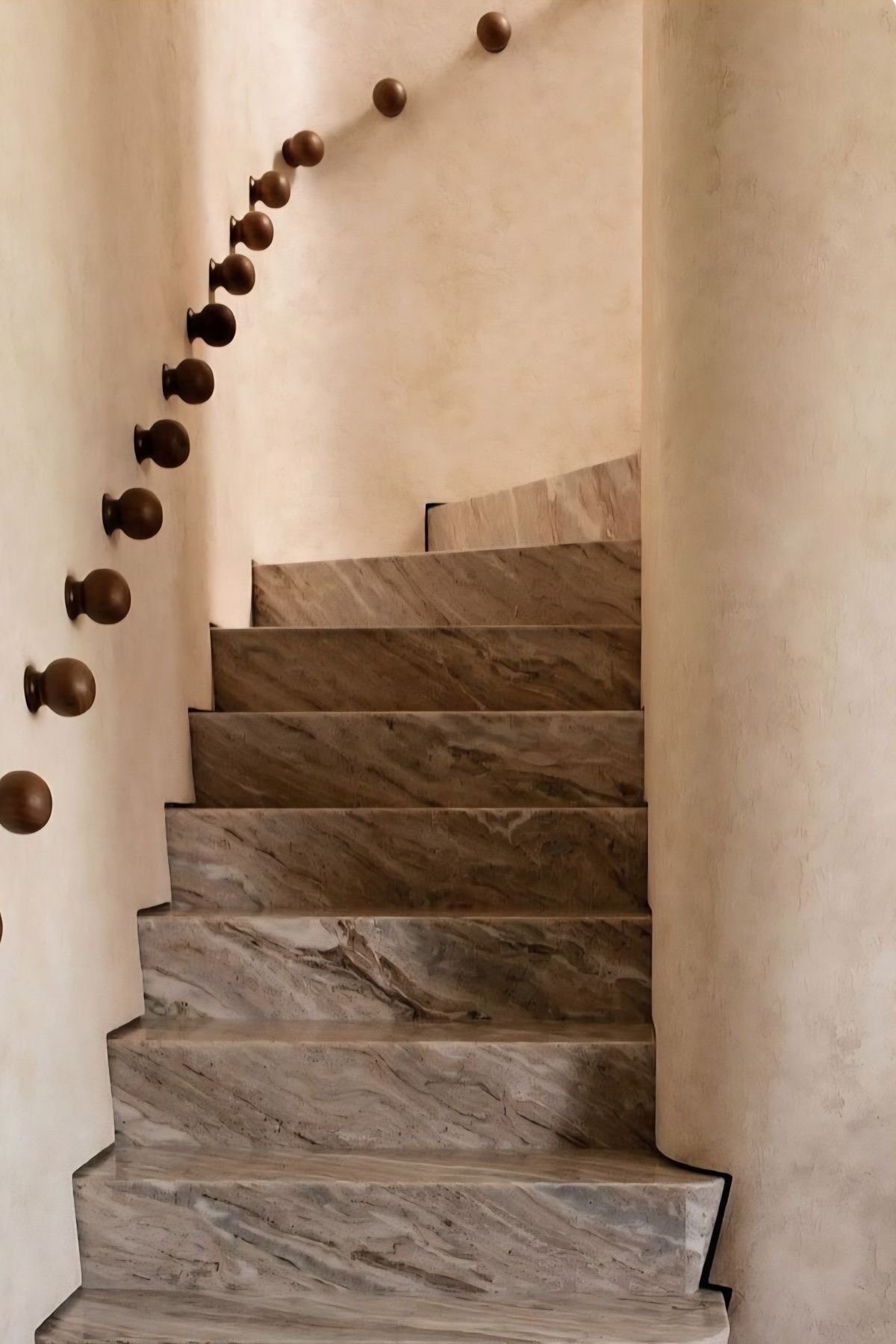 A staircase with marble steps and a curved beige wall on the right side. To the left, there is a handrail with round wooden balusters mounted on the wall.