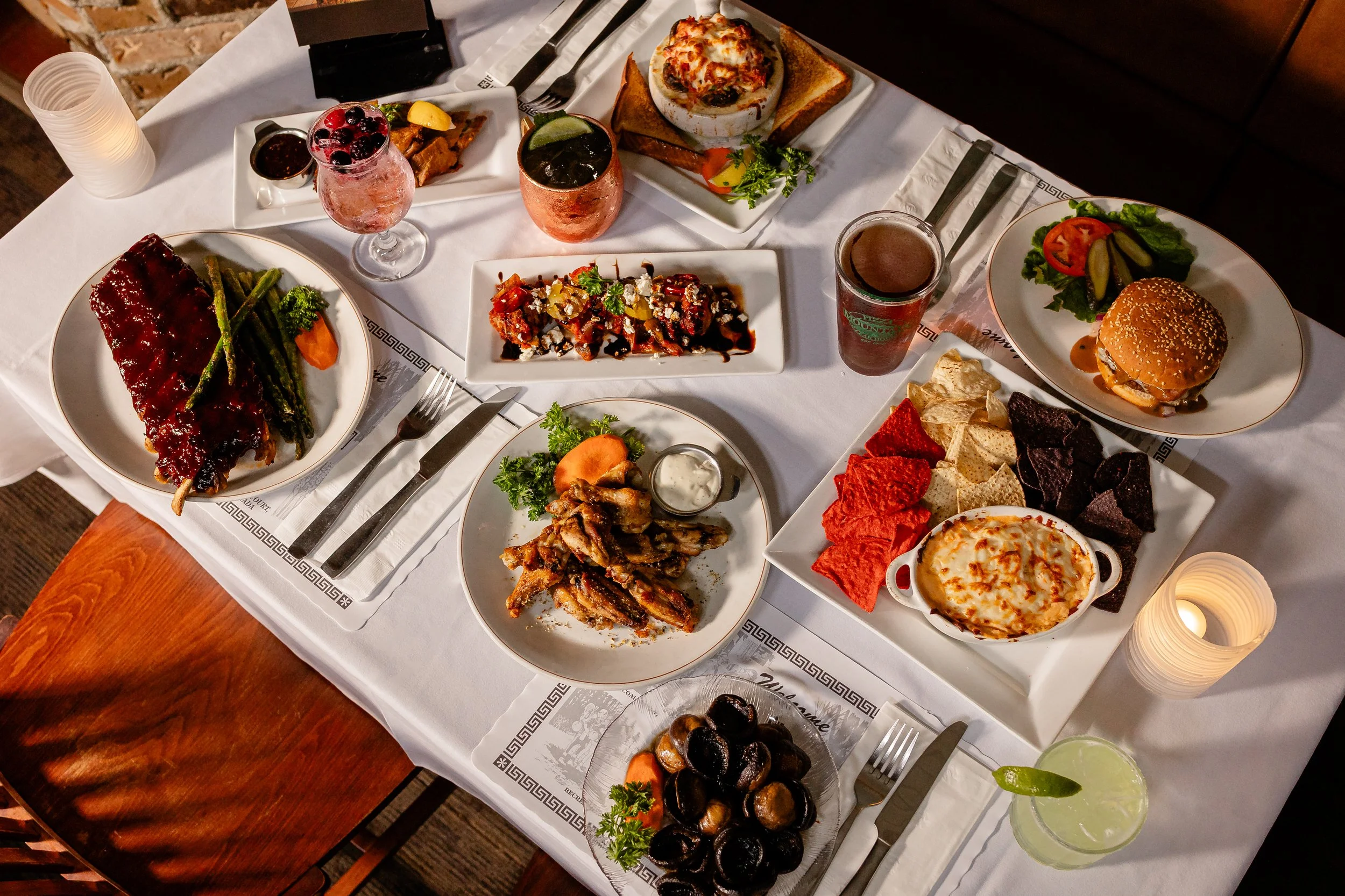 A table set with various dishes including a large ribs with green beans and carrots, a pizza, a salad with tomato and pickles, a cheeseburger, a dish with various toppings, a bowl of roasted mushrooms, a plate of nachos, and drinks, all on a white ta