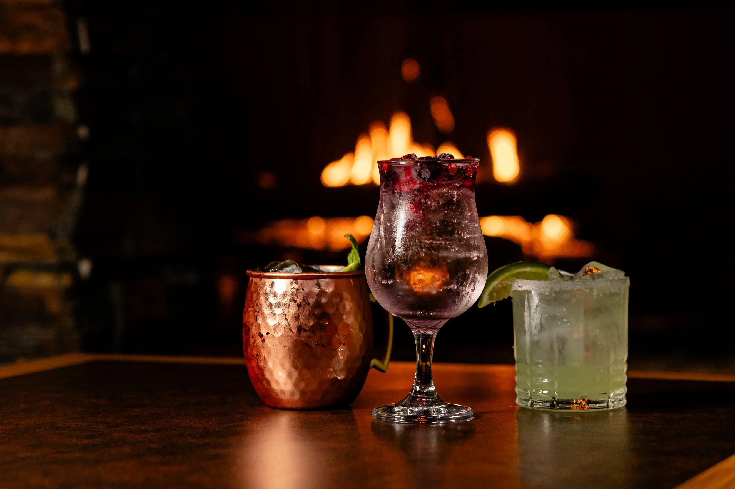 A close-up of a cocktail glass with ice and berries, with a glowing fire in the background
