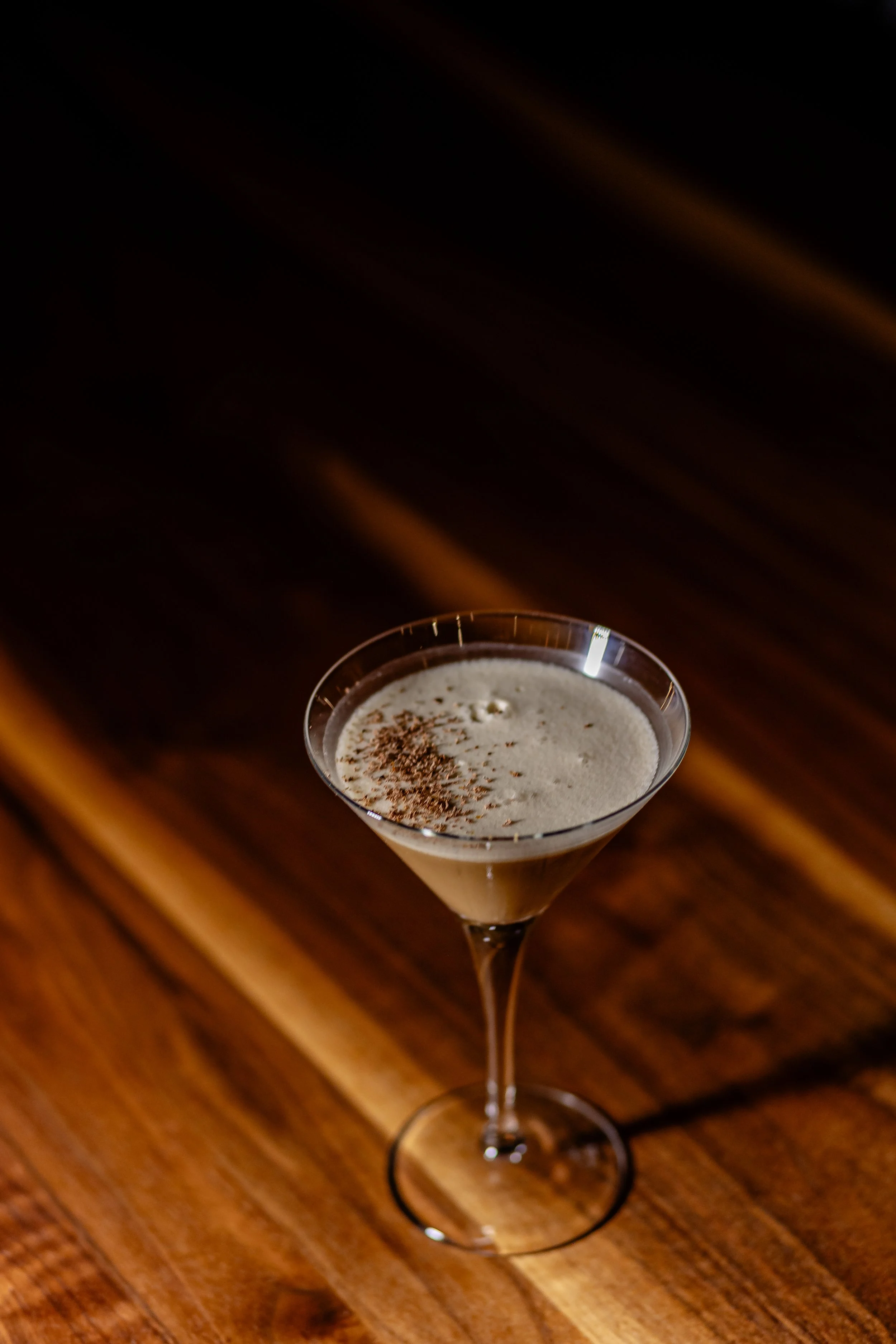 A elegant martini glass filled with a creamy coffee-flavored cocktail topped with chocolate shavings, placed on a wooden surface.