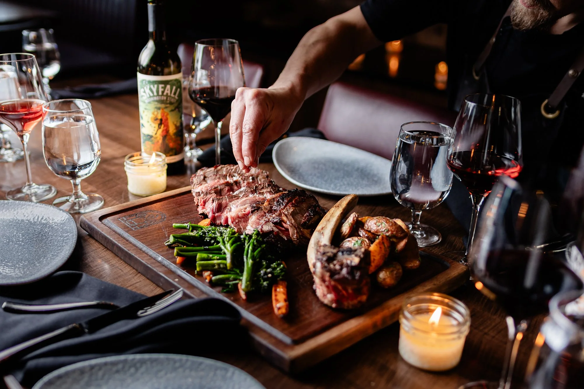 A person is serving a sliced roasted piece of meat on a wooden platter along with green beans and roasted vegetables. There are several glasses of red and white wine, a bottle of SkyFall wine, and lit candles on the table, creating an elegant dining 