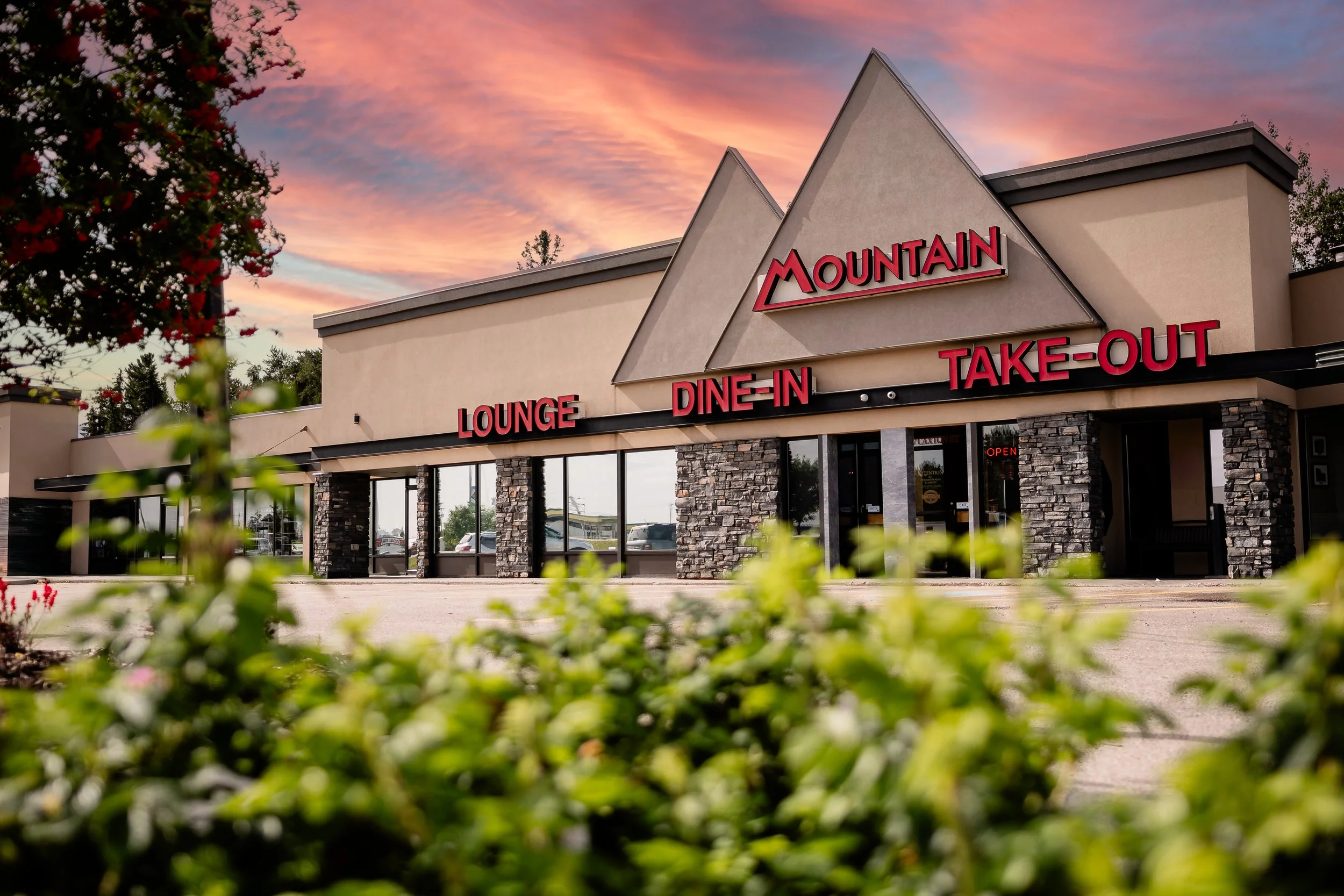 Exterior of a restaurant named 'Mountain' with signage indicating lounge, dine-in, and take-out options, during sunset with a colorful sky in the background.