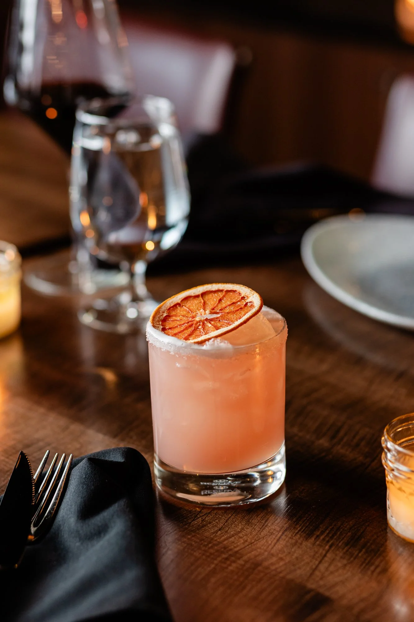 A pink cocktail with a dried orange slice garnishing the rim, placed on a wooden table at a restaurant.