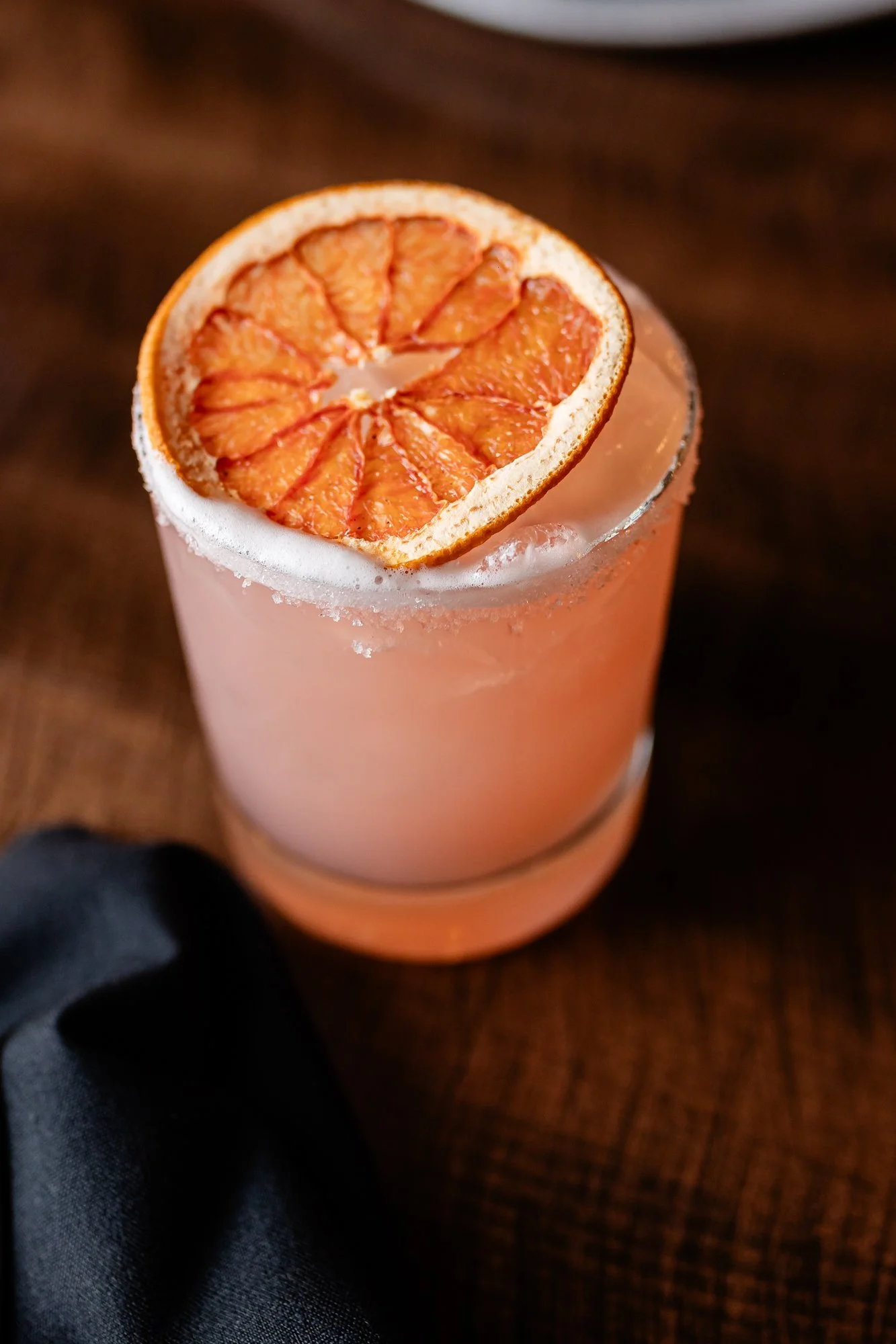 Close-up of a pink grapefruit cocktail in a clear glass, garnished with a dried grapefruit slice, on a dark wooden surface.