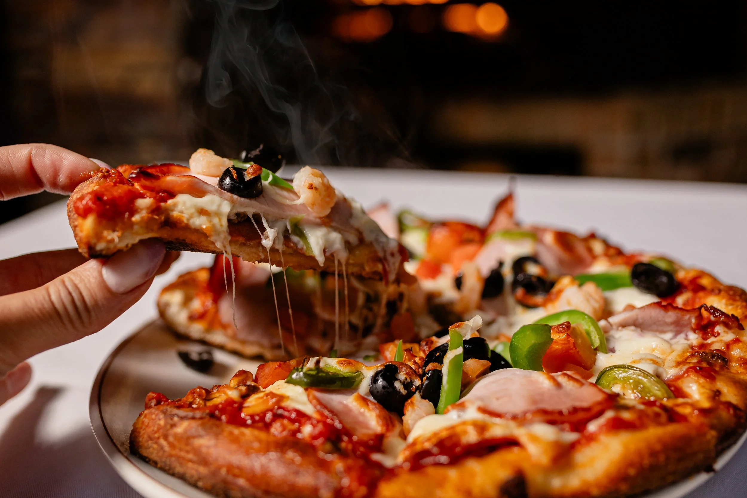 Close-up of a person holding a slice of pizza with melted cheese, toppings including black olives, shrimp, and green bell peppers, above a pizza with various toppings on a plate.