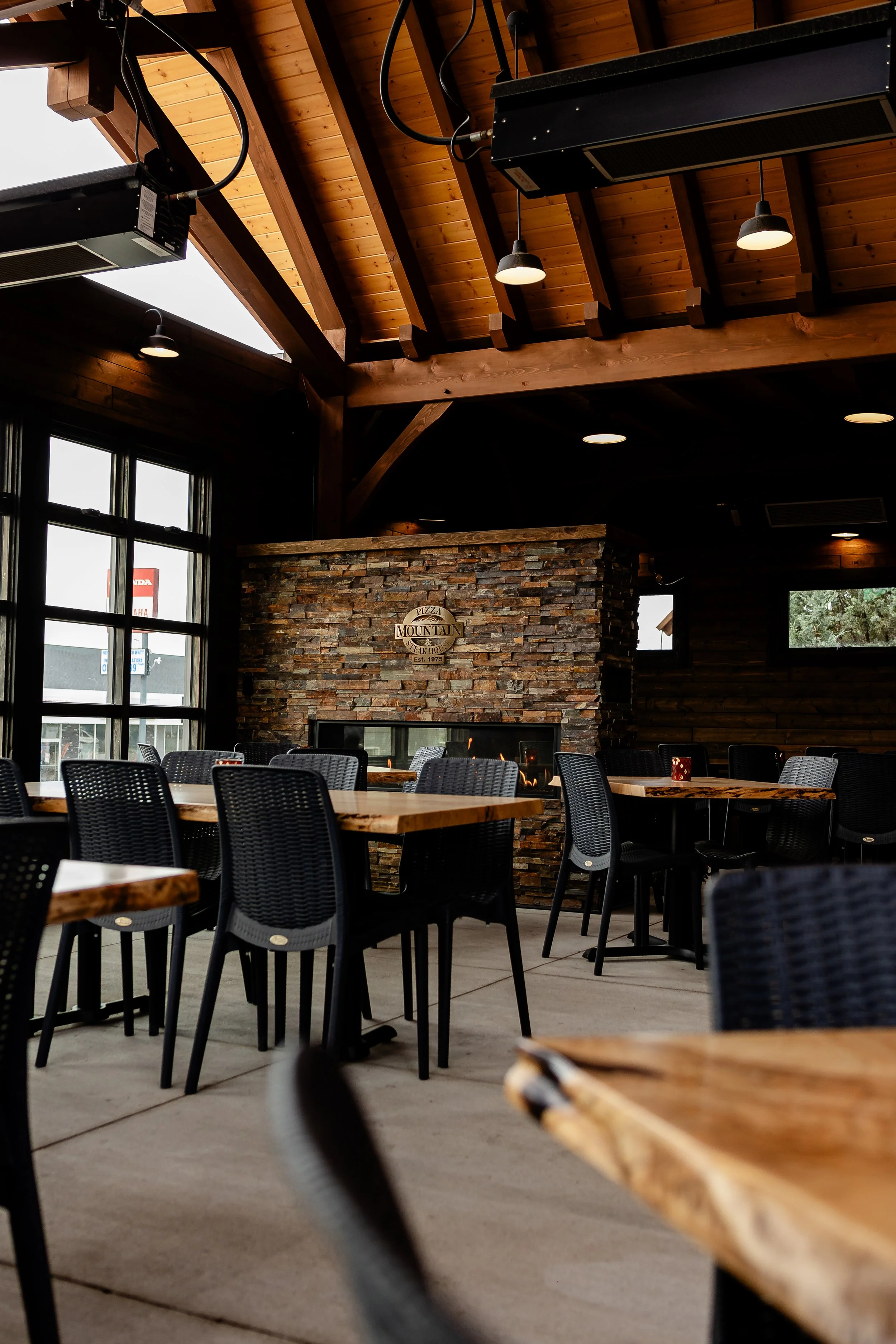 Inside a restaurant with wooden ceiling beams, a stone fireplace, and black chairs around wooden tables. Large windows let in natural light.