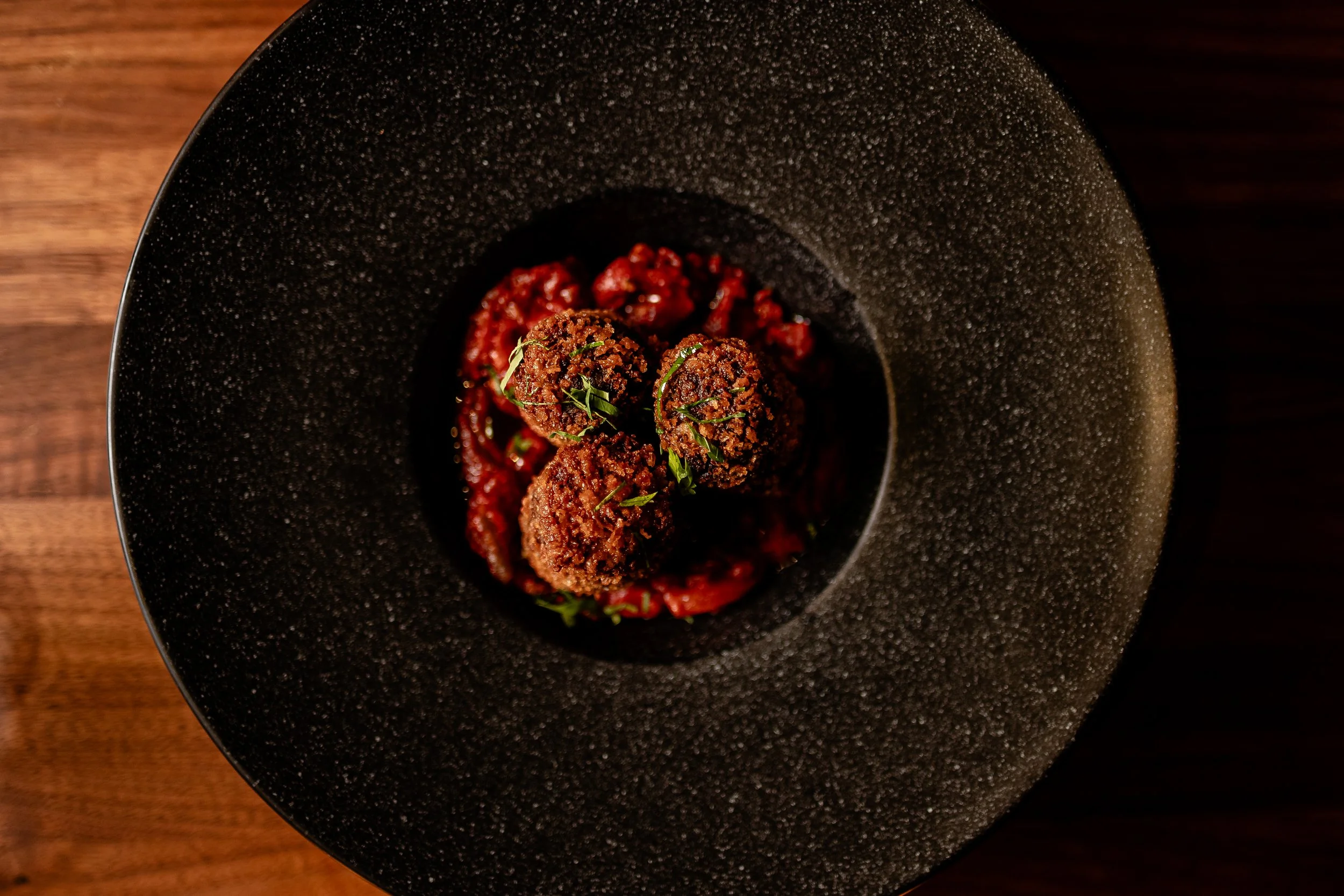 Close-up of a textured black bowl with three falafel balls and red sauce, garnished with chopped herbs, on a wooden surface.