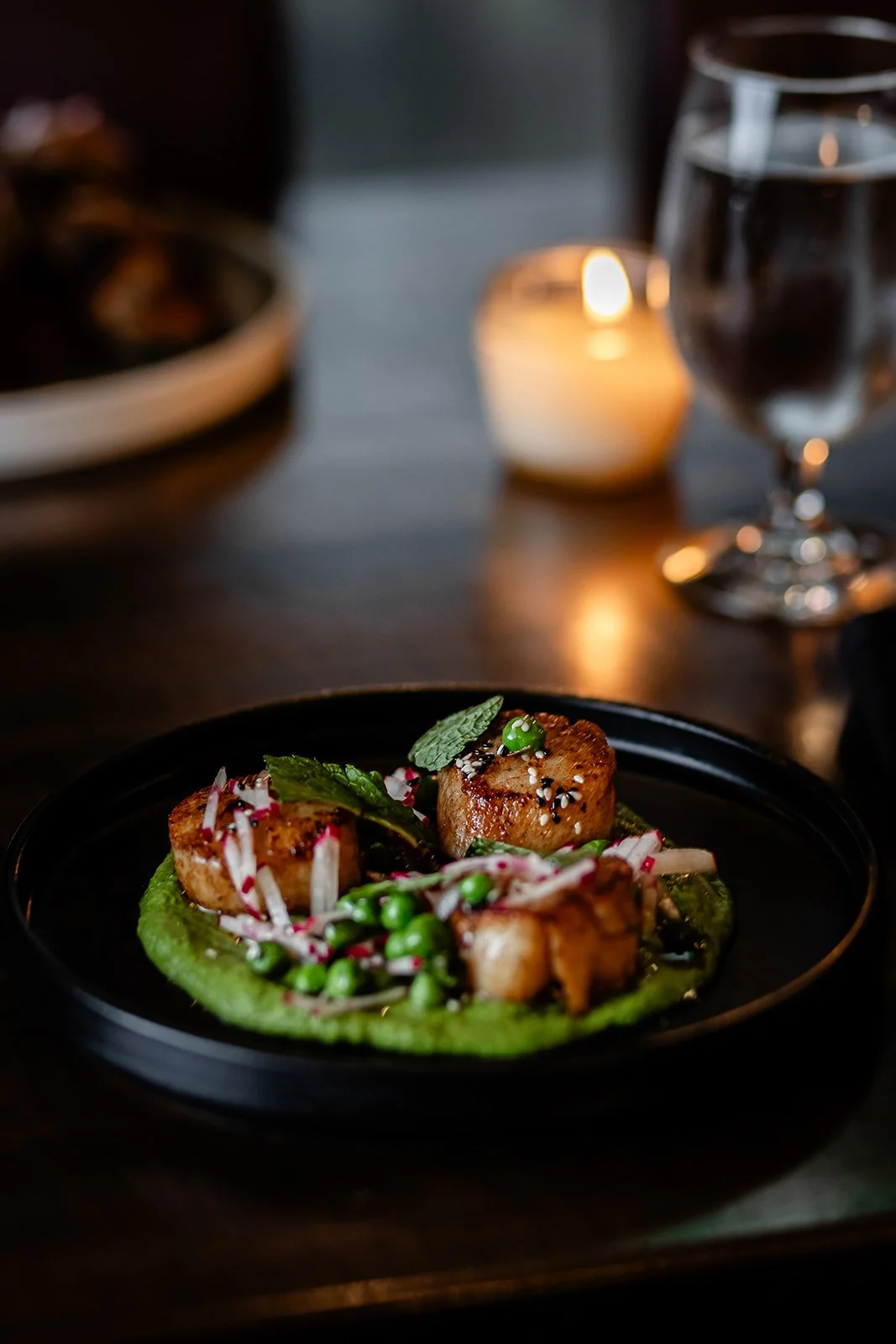 Close-up of a gourmet dish with seared scallops, green peas, radish slices, and mint leaves on a bed of green sauce, served in a black bowl on a dark table with a water glass, candle, and blurred background.