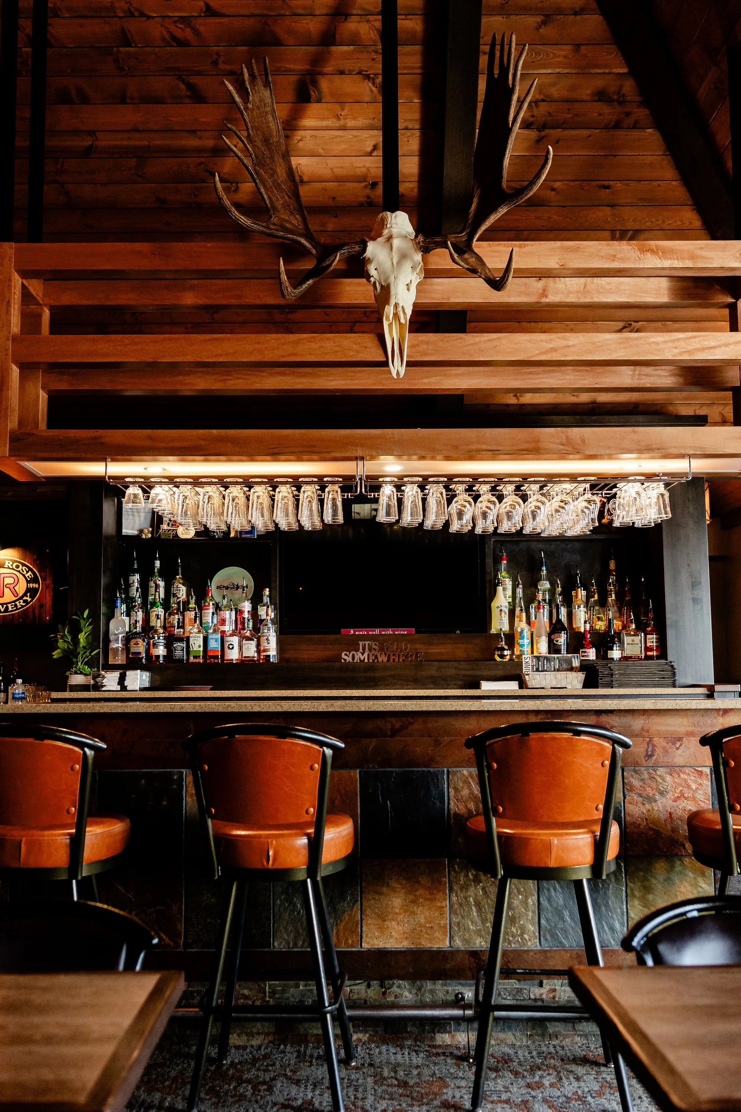Interior of a bar with a wooden ceiling, bar counter with various bottles of alcohol, hanging glassware, and leather bar stools with black legs and backs.