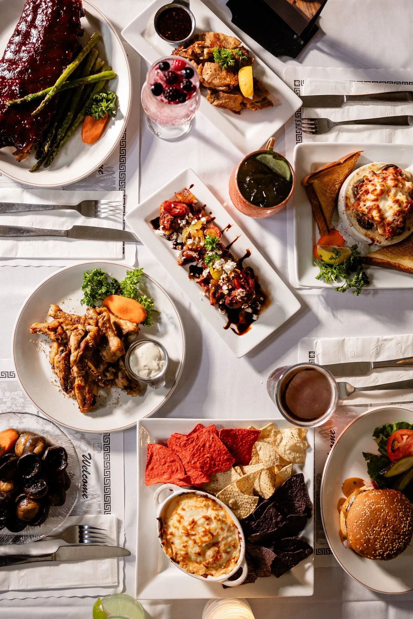 A table set with various dishes of food, including a burger, a baked dish with cheese, chips with salsa, stuffed grape leaves, a dish with meat and vegetables, a milkshake with berries, a dark soda, chips with dip, and a dessert with caramelized cheese, all arranged on white plates and a white tablecloth.