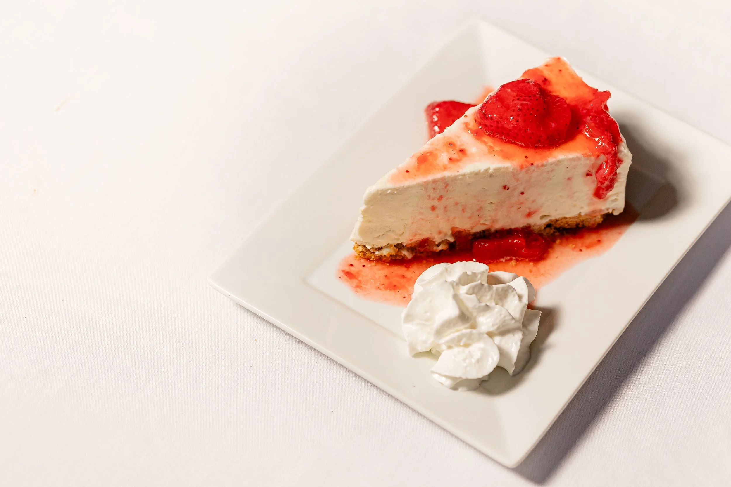 Slice of strawberry cheesecake on a white plate, topped with fresh strawberries and a drizzle of strawberry sauce, served with whipped cream.