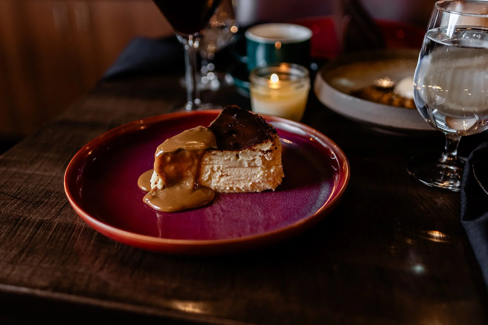 A slice of cheesecake topped with caramel sauce on a red plate, set on a dark wooden table with a glass of water, a glass of red wine, a small candle, and plates in the background.