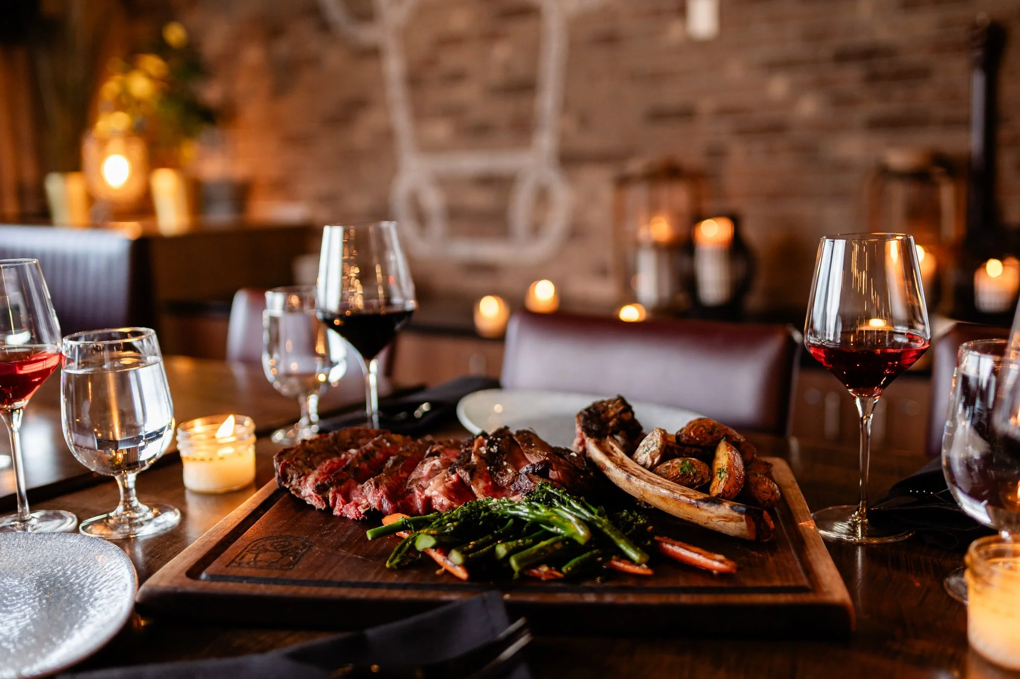 A table set for dinner with glasses of red wine, water, and candles, featuring a platter of ribs, grilled vegetables, and asparagus in a cozy, dimly lit restaurant with a brick wall background.