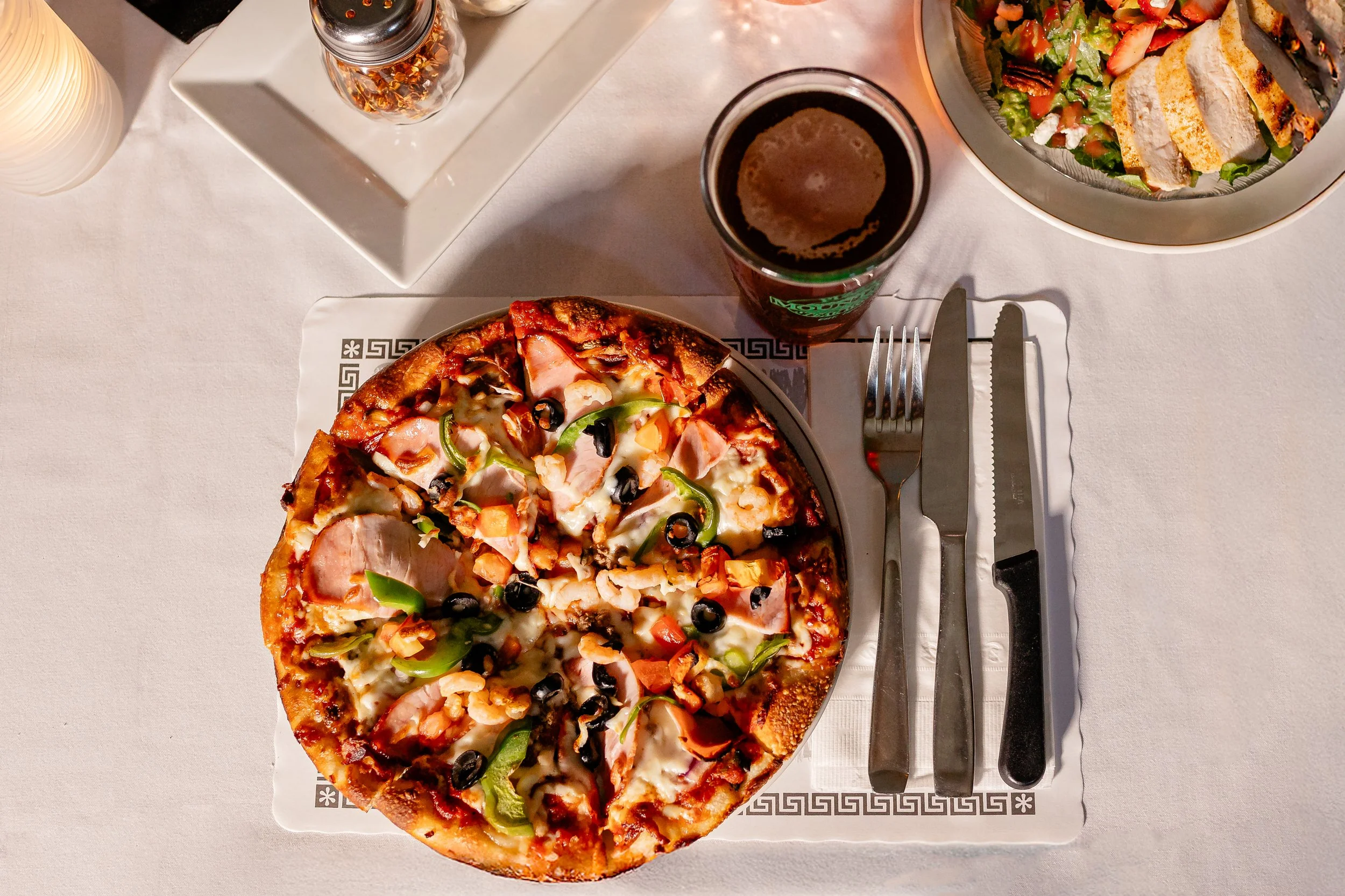 Pizza with various toppings including black olives, green peppers, and meats on a white plate. A salad with strawberries and greens in a bowl, a glass of dark soda, a napkin, and utensils on a white tablecloth.