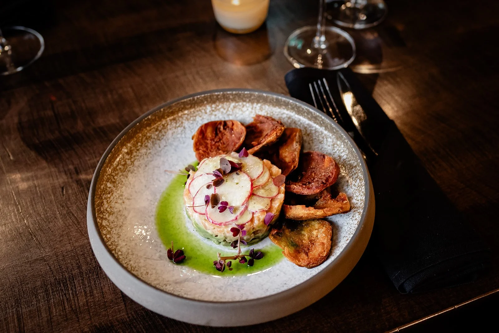 A white and gray ceramic plate with a layered salad topped with sliced radishes and microgreens, served with grilled vegetables like eggplant and zucchini, on a dark wooden table with wine glasses in the background.