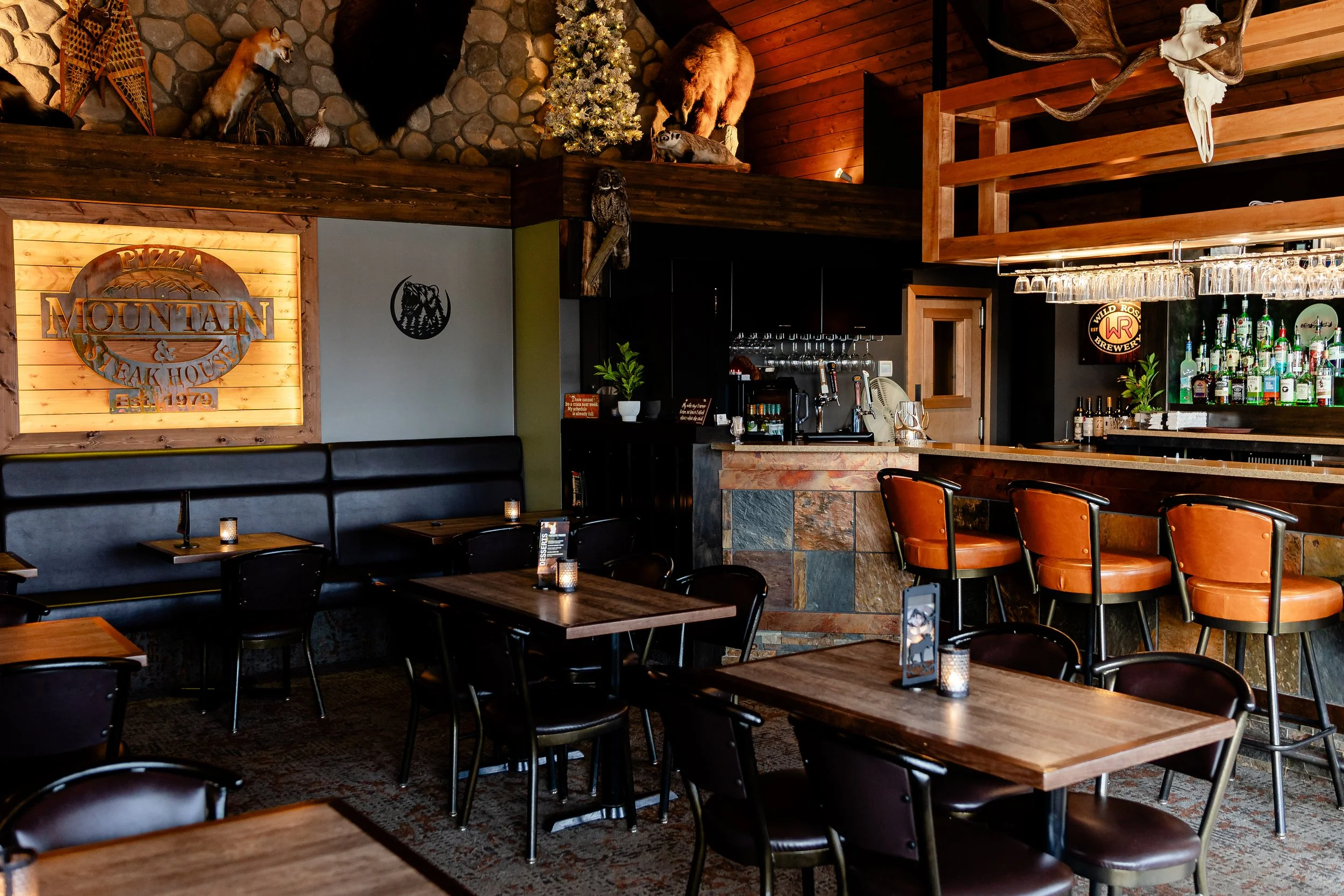Interior of a rustic restaurant or bar with wooden furniture, a bar counter with glassware, and themed decor including mounted animal heads and a Christmas tree.