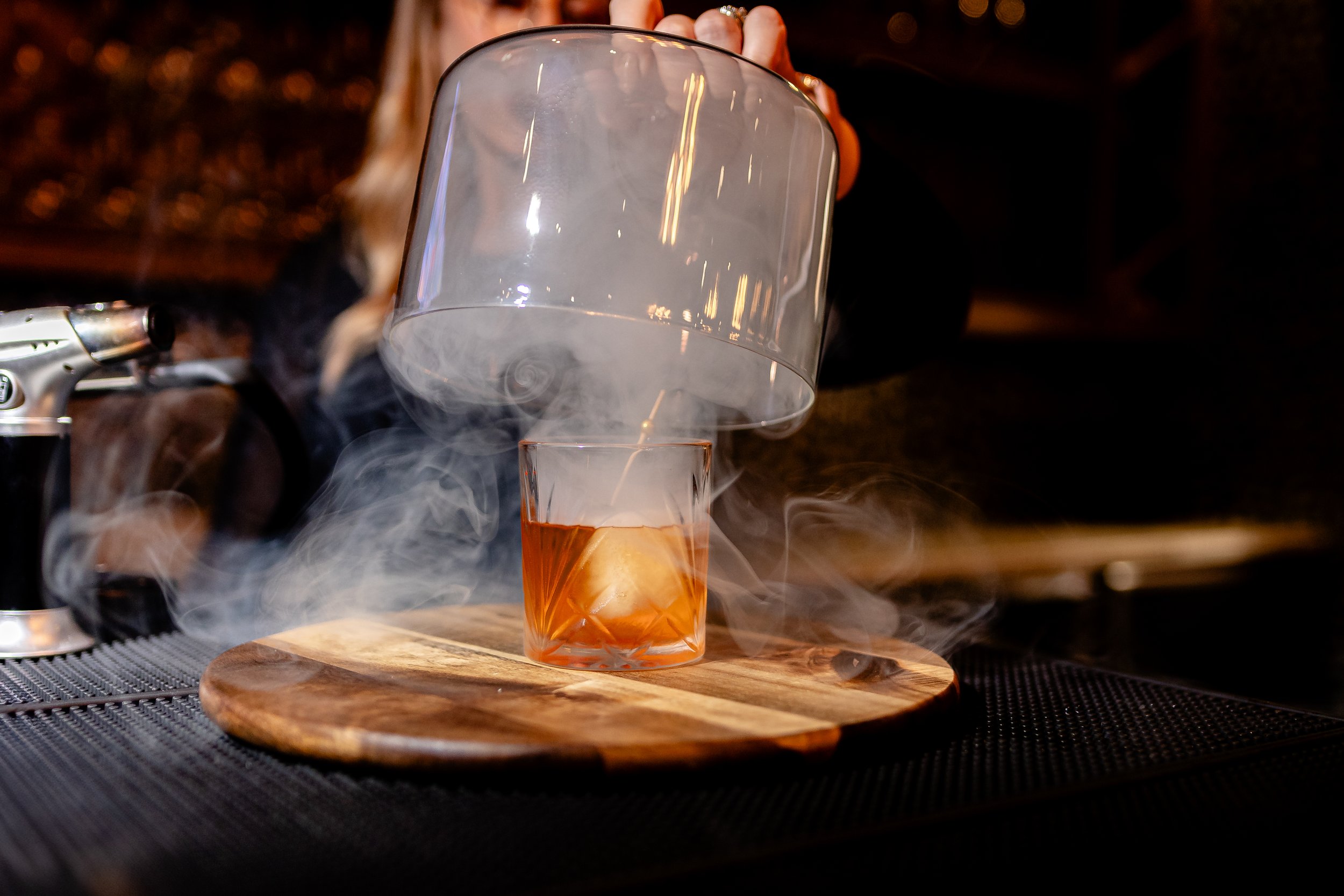 A bartender is pouring a drink into a glass. The glass is tilted and smoke or vapor is swirling around it, creating a smoky effect. The background is dark with warm lighting reflections.