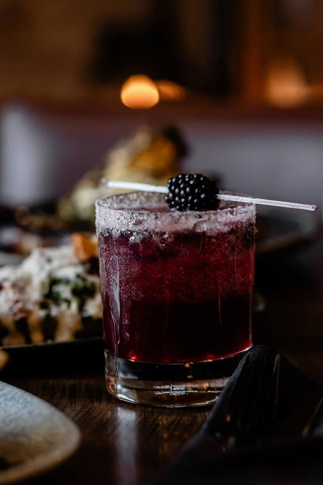 A close-up of a dark red cocktail in a short glass garnished with a blackberry on a clear skewer.