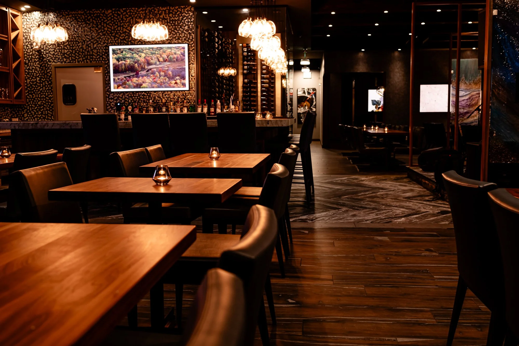 An elegant dimly lit bar area with a curved marble countertop, black bar stools, and bottles of alcohol displayed behind the bar. There are framed pictures and decorative lighting fixtures on the walls, along with wooden tables and chairs in the fore