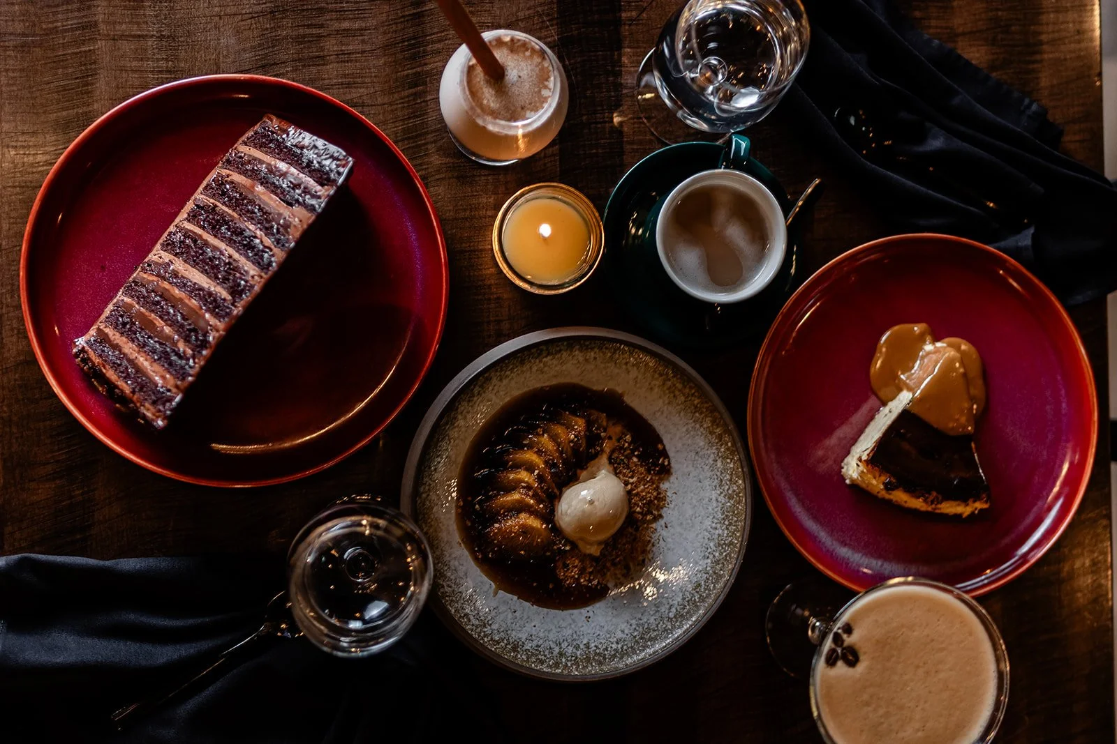Top-down view of a wooden table with various desserts, drinks, and a candle. There are two red plates with cake slices, one with caramel topping and the other with a layered chocolate and caramel cake. A small dish with a pastry topped with sauce and a scoop of ice cream, a glass of water, a coffee cup, and several coffee beverages are also visible. The setup is cozy and elegant.