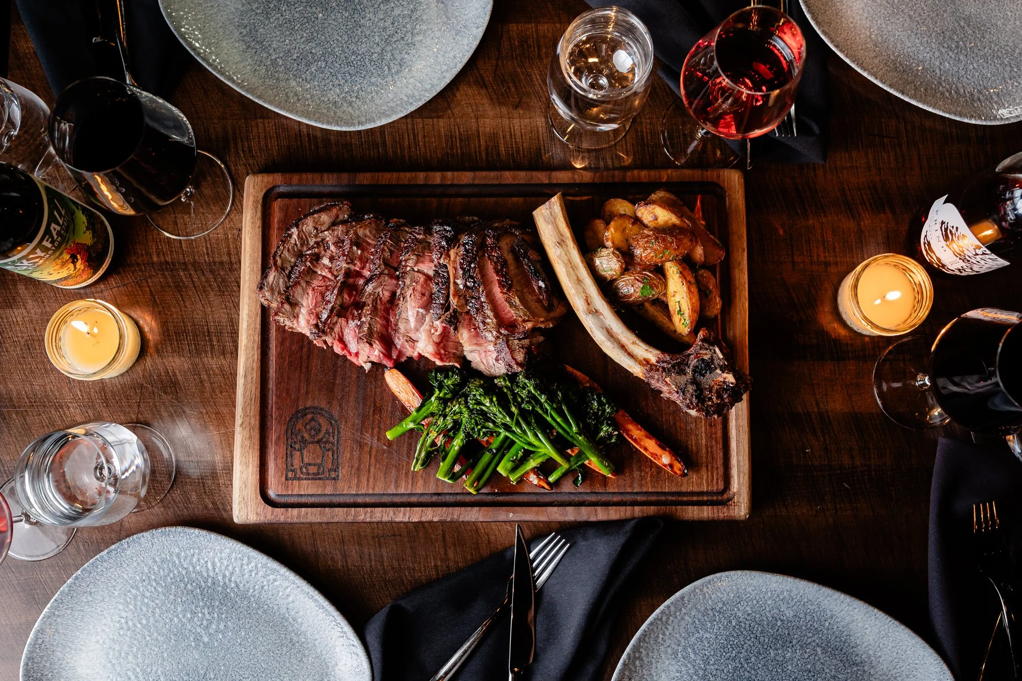 A wooden table with a large grilled steak with grill marks, served with roasted potatoes, carrots, and green vegetables. Surrounded by several glasses of red wine, water, and wine bottles, with votive candles and black napkins on the table.