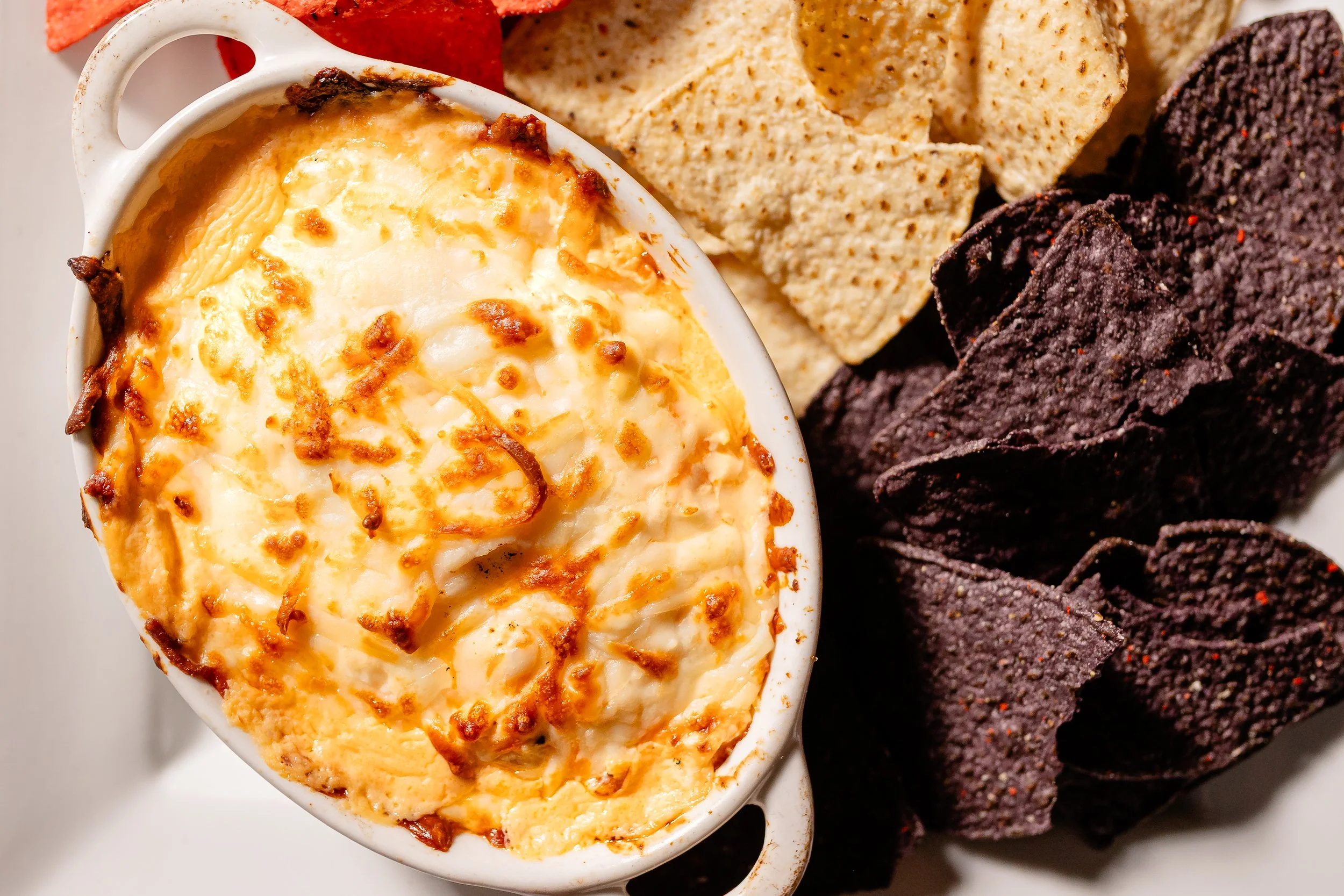 Close-up of a baked dish topped with melted cheese in small white ramekins and a bed of assorted tortilla chips on the side.