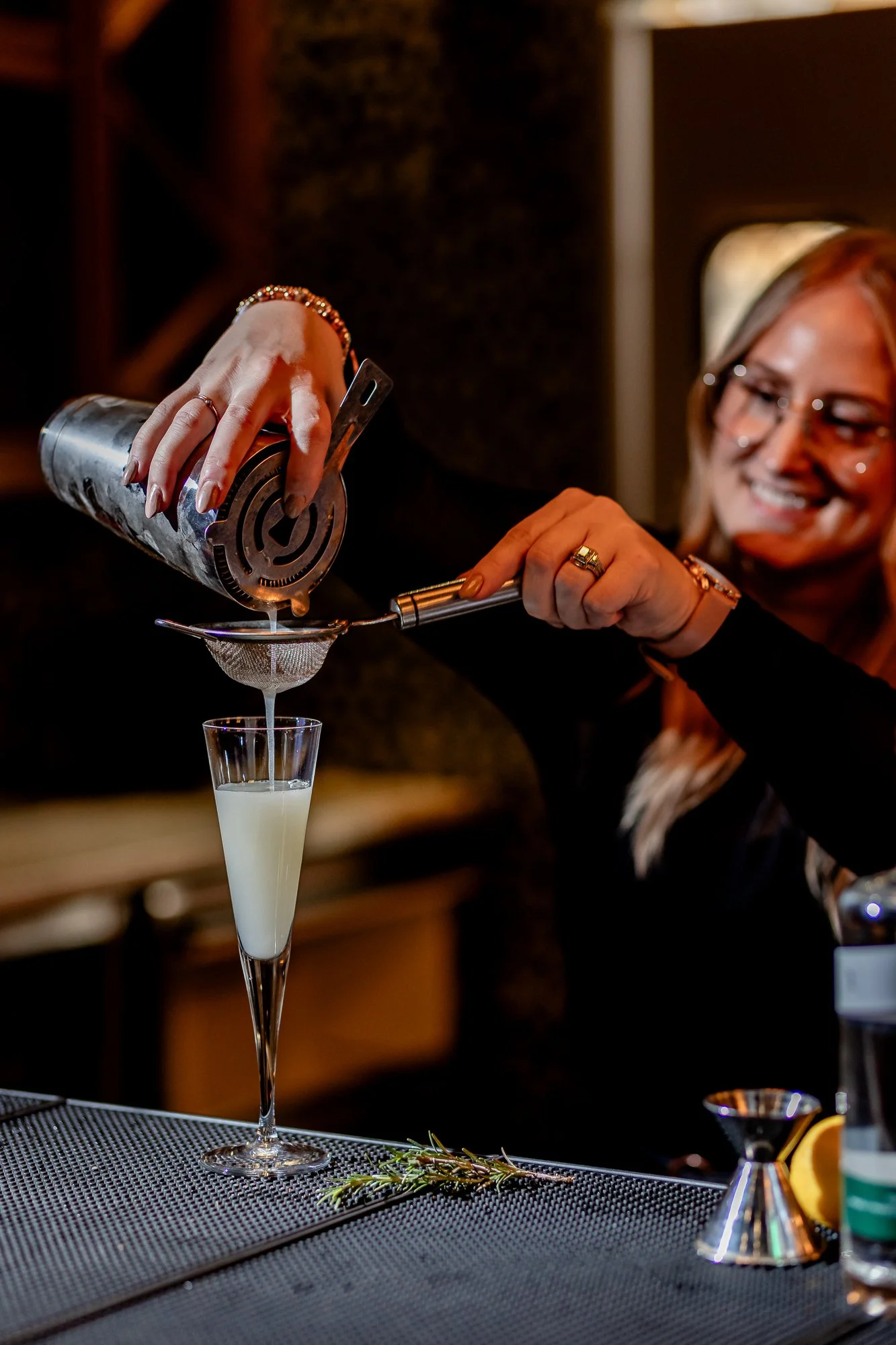 A woman with blonde hair and glasses is smiling while pouring a white liquid from a cocktail shaker into a tall champagne flute through a strainer, with a sprig of rosemary and lemon slices on the bar in the foreground.