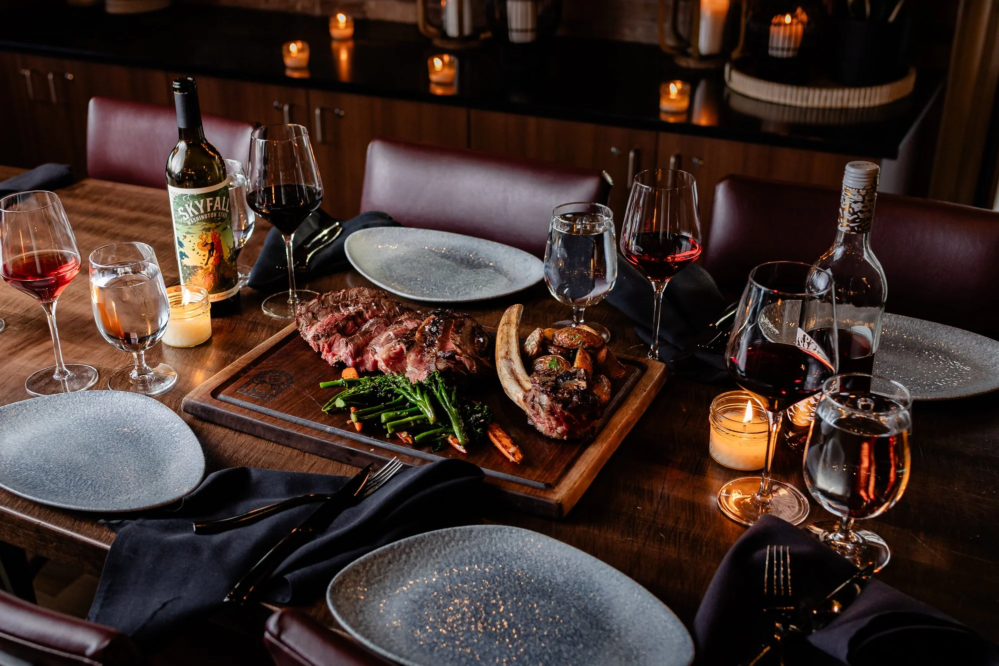 A dinner table set with wine glasses, a bottle of red wine, a steak with green vegetables, a rib bone, and empty plates. Candlelight and candles create a warm ambiance in a dimly lit dining room.