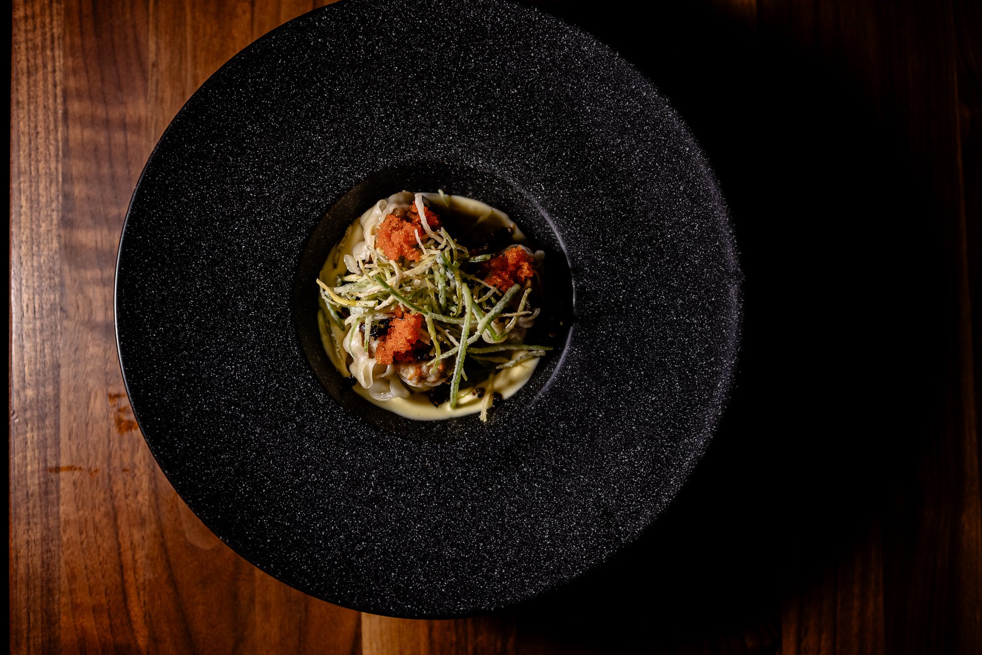 A black, textured plate with a pasta dish topped with green herbs, red-orange sauce, and grated cheese, on a wooden table.