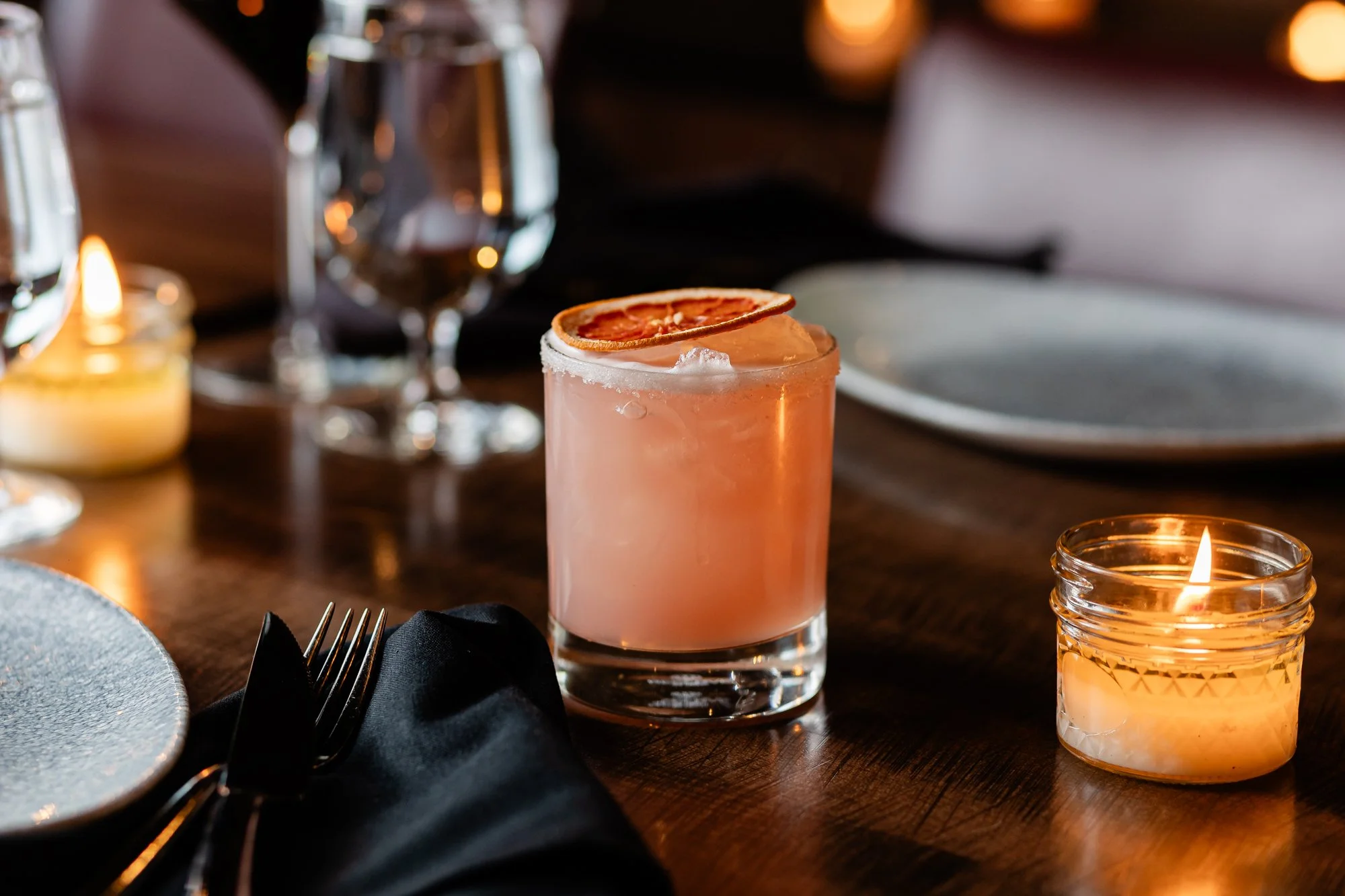 A pink cocktail with a dried orange slice garnish on a wooden dining table, surrounded by candles and glassware, with a blurred background of a formal dinner setting.