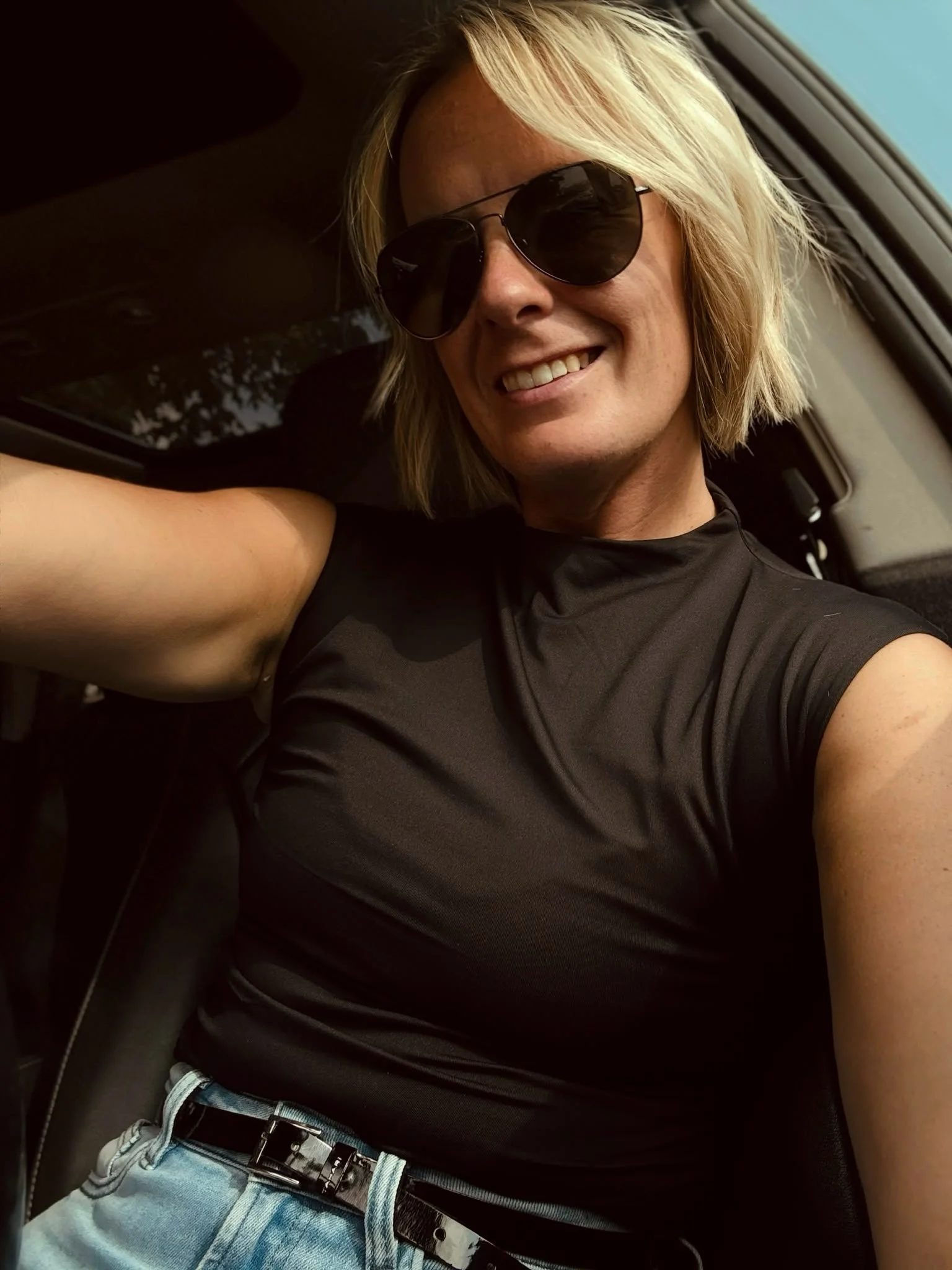 A person with blonde hair wearing black sunglasses, a black shirt, and light blue jeans sitting in a vehicle, smiling and taking a selfie.