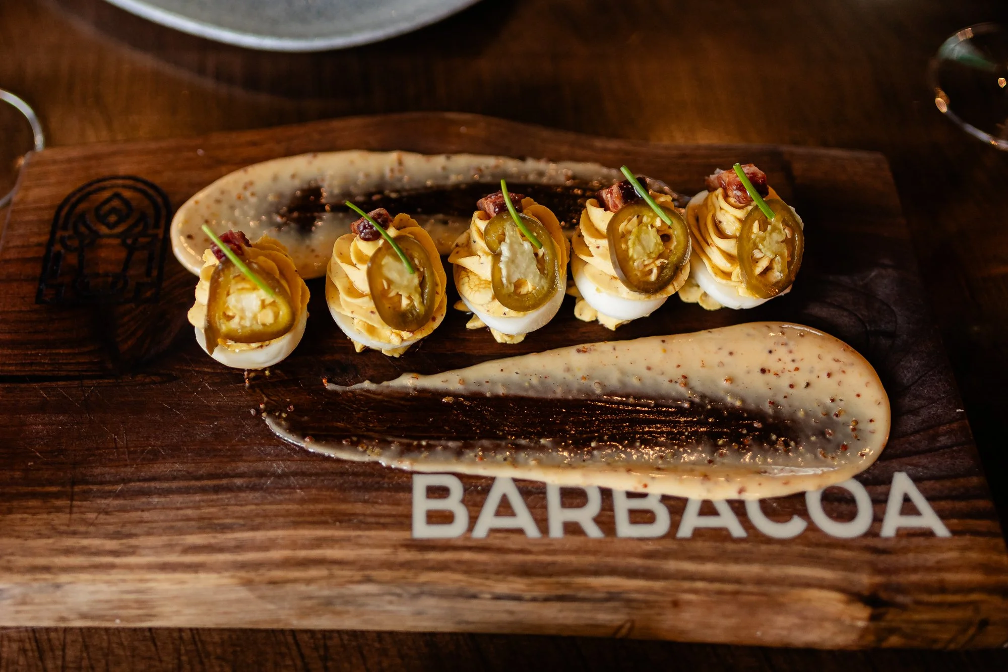 Deviled eggs garnished with jalapeño slices, bacon pieces, and chives, served on a wooden board with a drizzle of sauce.