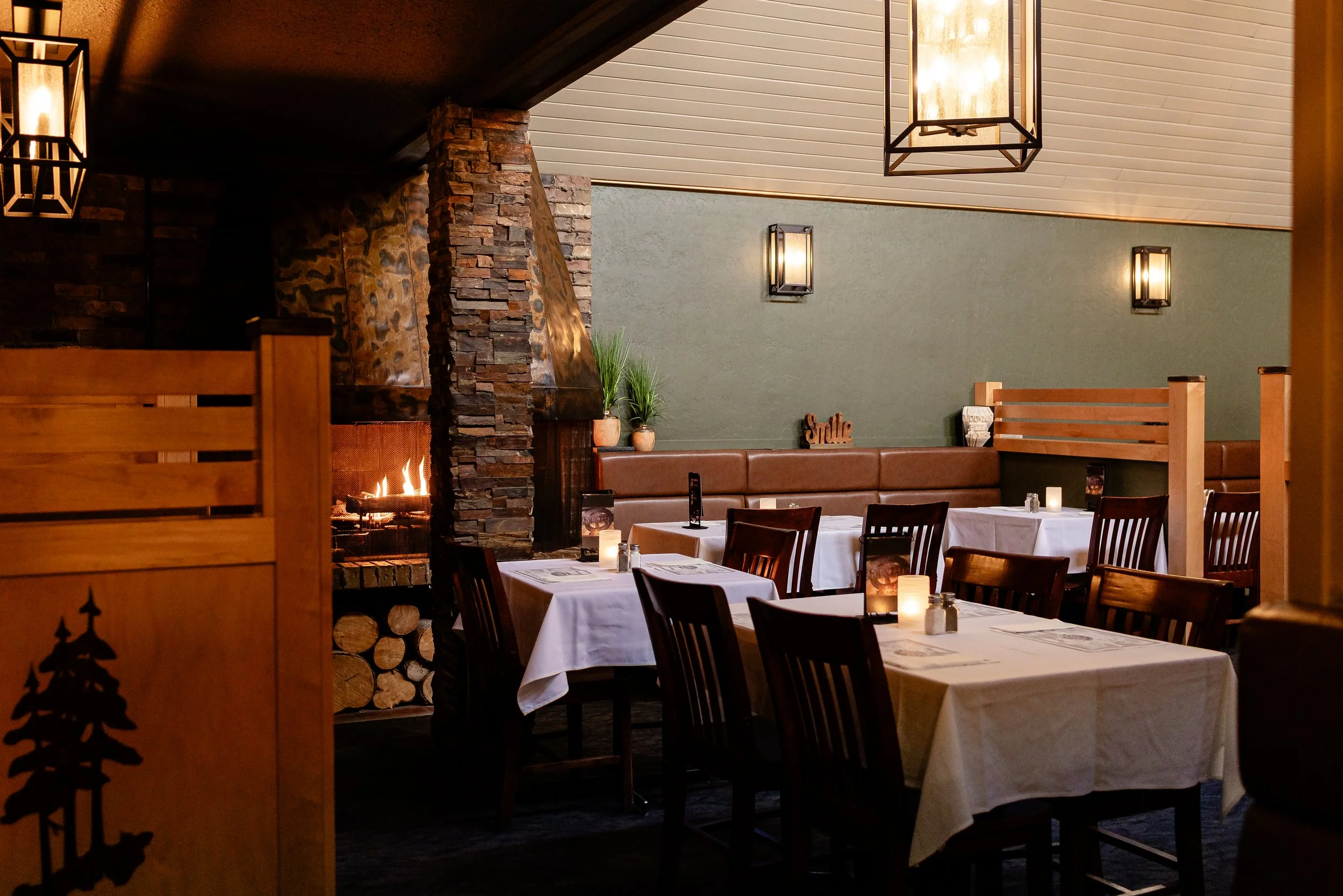 Inside a cozy restaurant or dining area with tables covered in white tablecloths, lit candles, and wooden chairs. There is a stone fireplace, potted plants, and wall-mounted lantern-style lights along a green wall. The ceiling has a decorative wooden