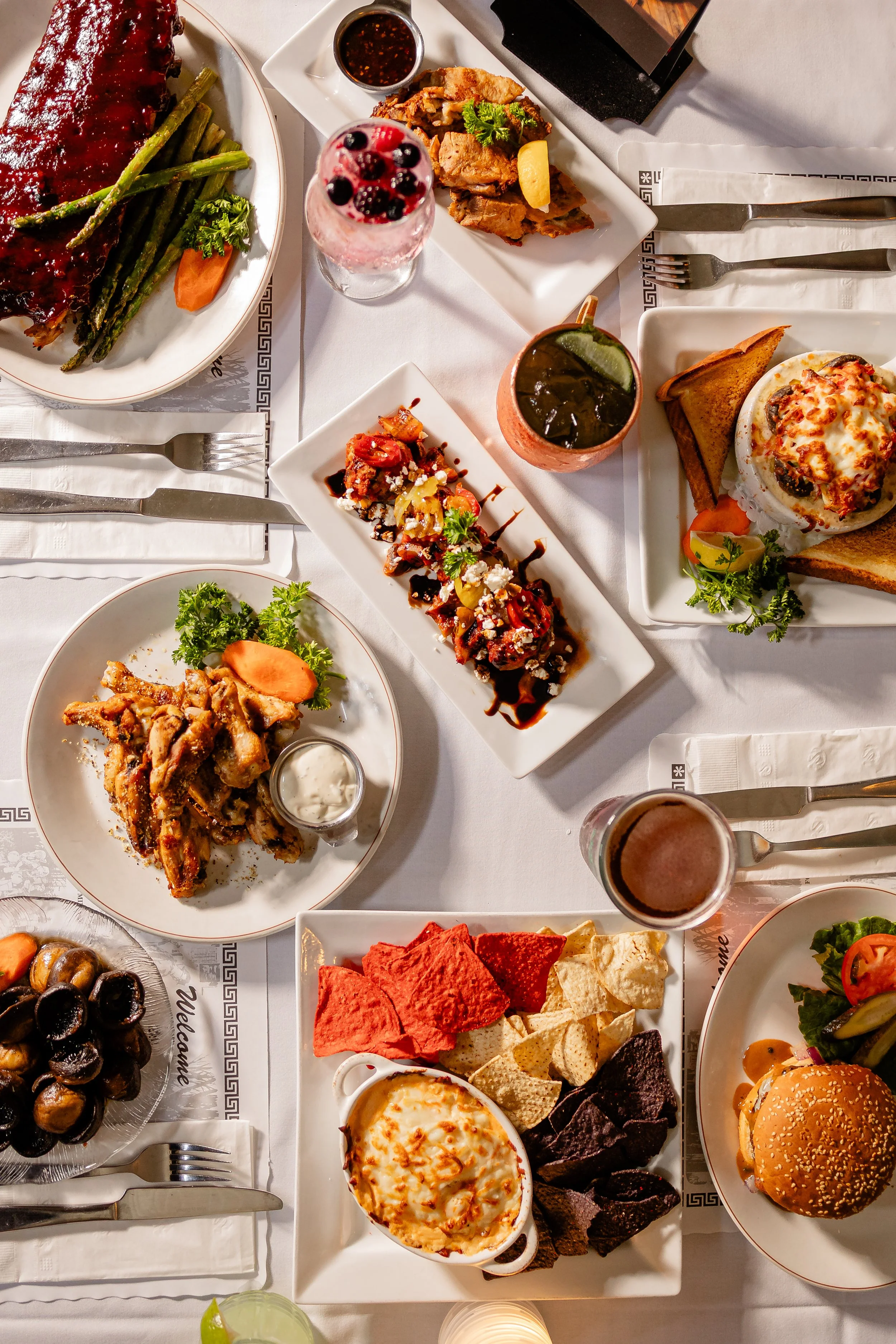 A variety of dishes on a white tablecloth at a restaurant, including a plate of ribs with green beans and a carrot, a dessert with berries and whipped cream, a salad with greens and cherry tomatoes, breaded chicken with a lemon wedge, potato wedges, 