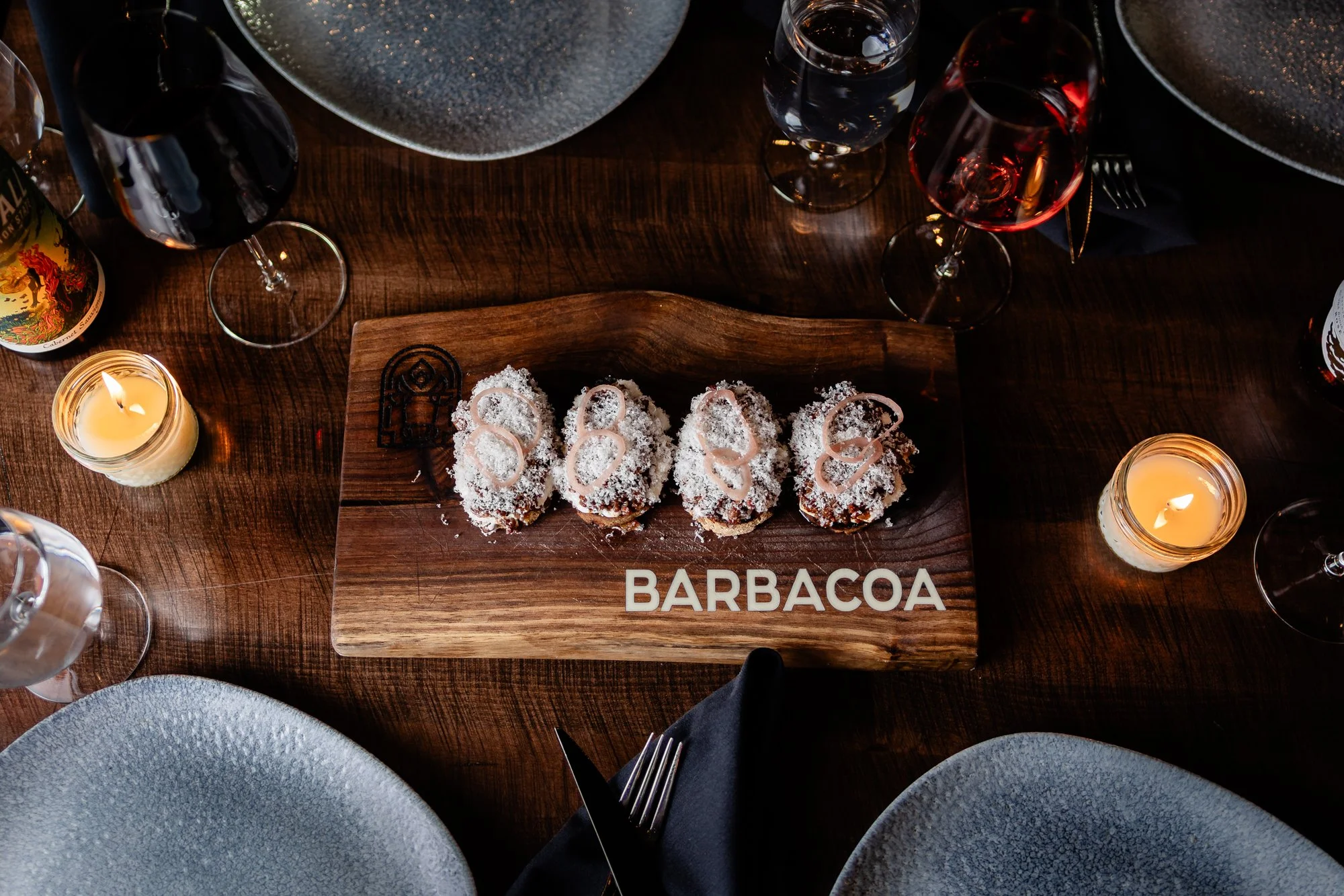 Top-down view of a dining table set with blue bowls, wine glasses, candles, and a wooden platter with four pieces of dessert labeled 'Barbacoas' in white letters.