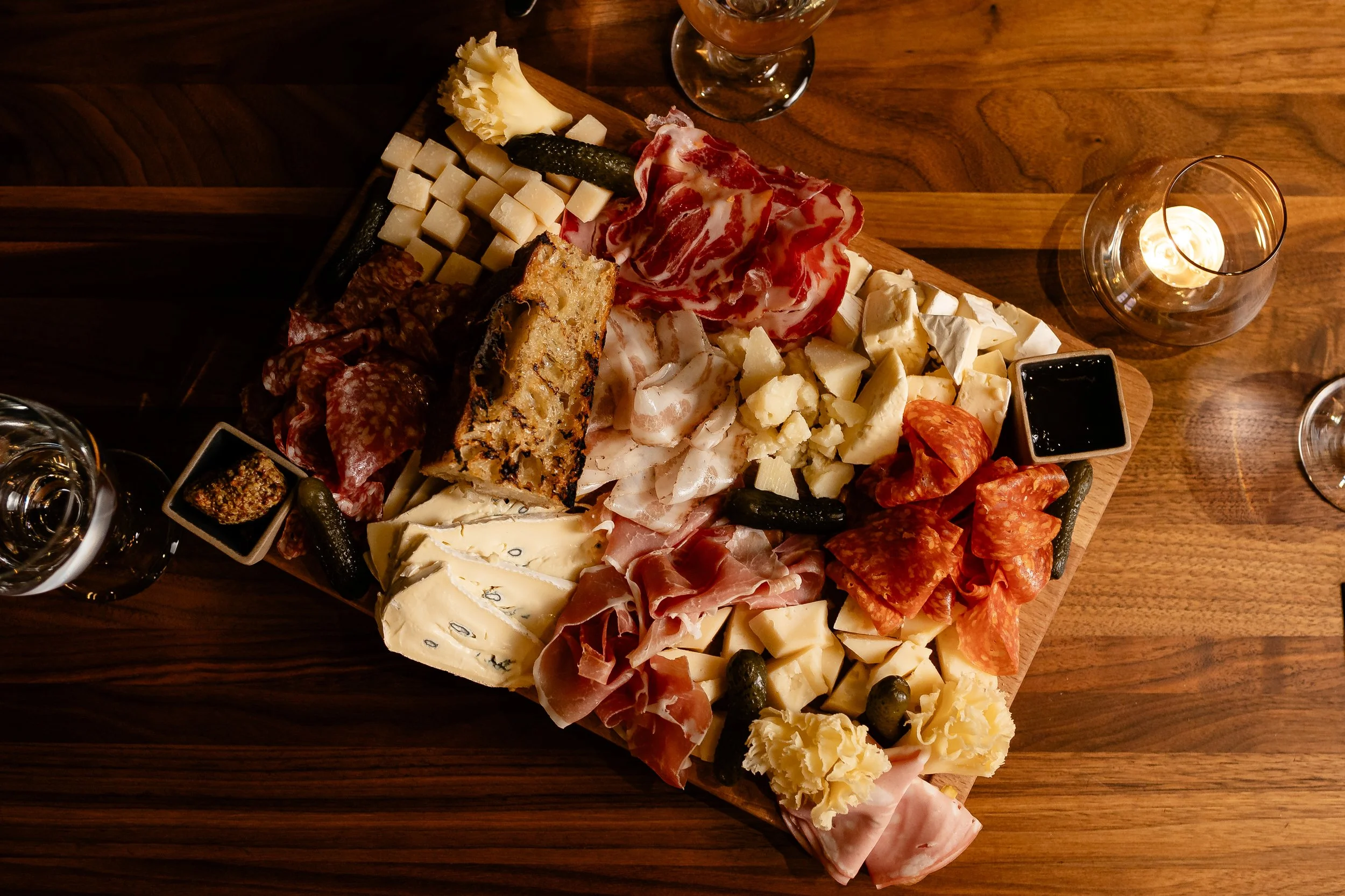 A wooden charcuterie board featuring an assortment of cheeses, cured meats, crackers, and pickles, with small cups of dark sauces and candles in glass holders for ambiance.
