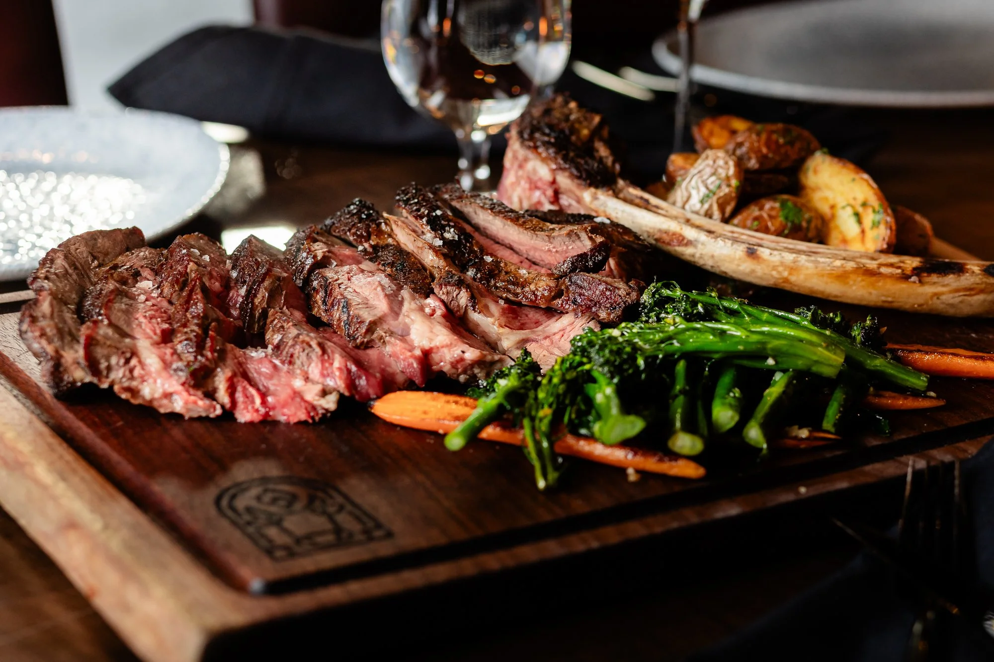 A wooden platter of grilled meats including sliced steak and rack of lamb, accompanied by green beans, carrots, and roasted potatoes.